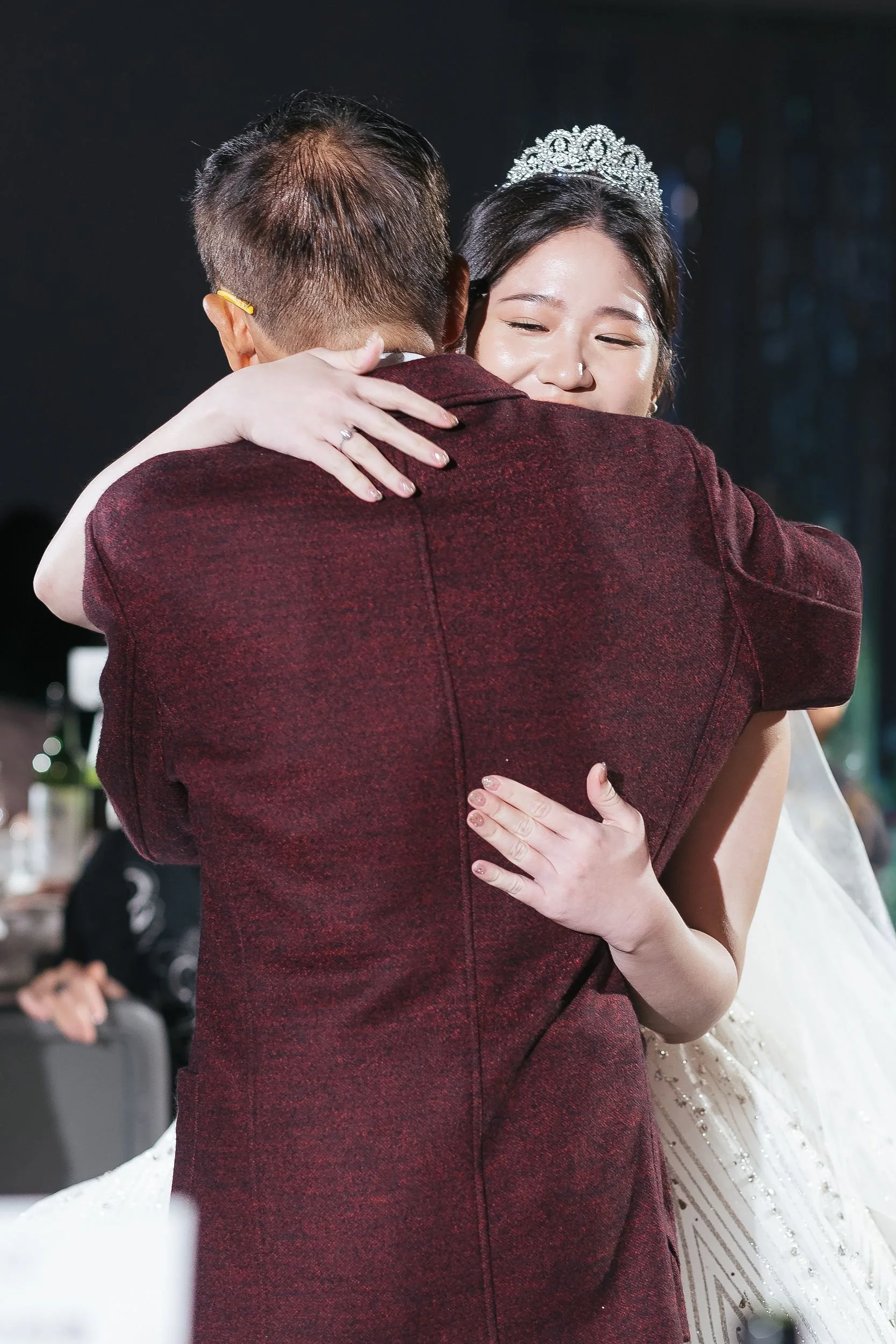 Young woman with a jeweled tiara hugging a man in a burgundy suit at a celebration or wedding.
