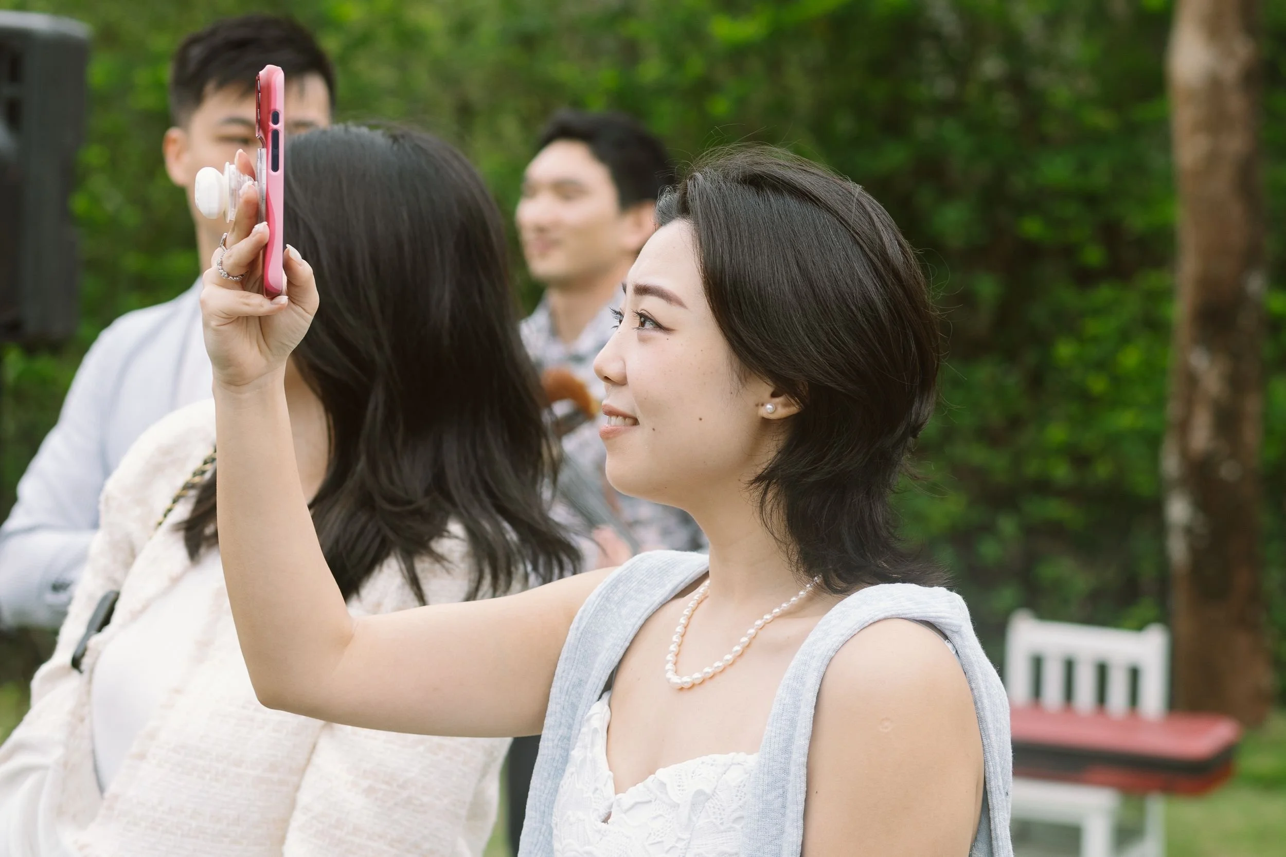 A woman takes a selfie with a group of people outdoors in a park with green trees in the background.