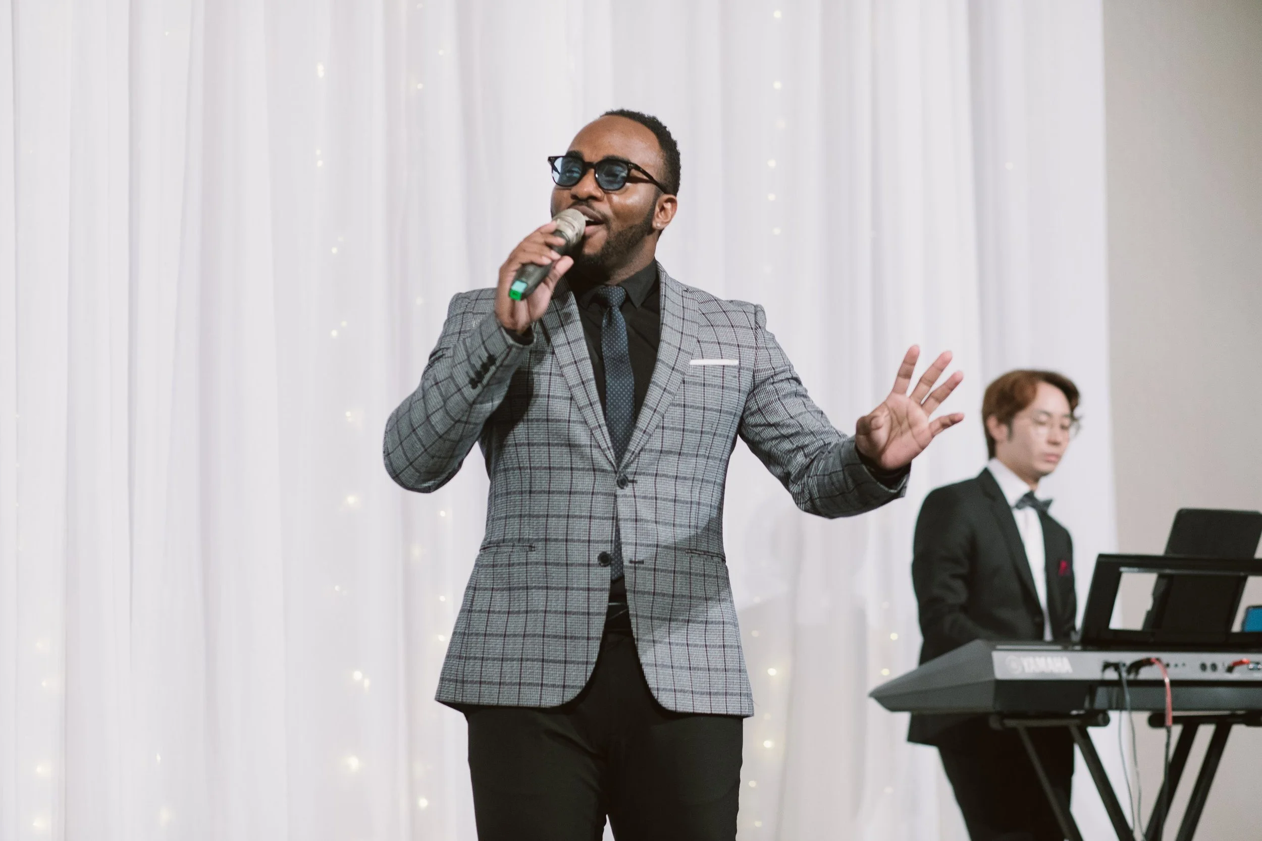 A man in a gray checkered suit with sunglasses singing into a microphone at an event with white draped background and musicians in the background.