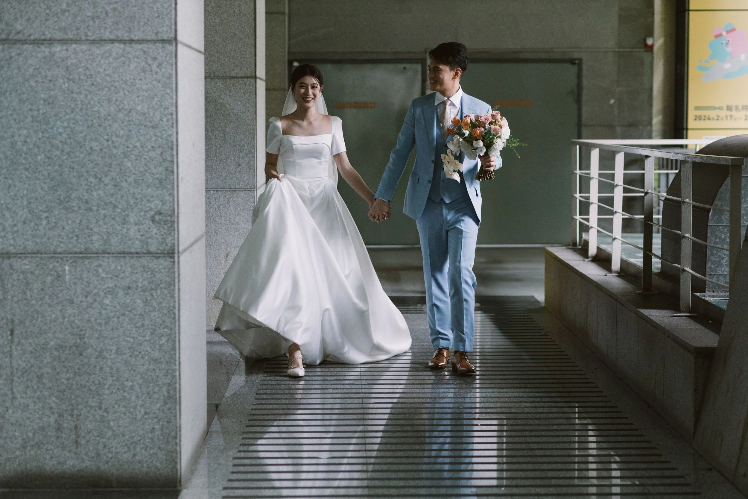 A bride and groom holding hands and walking indoors, smiling at each other. The bride is wearing a white wedding gown and veil, and the groom is in a light blue suit holding a bouquet of flowers.