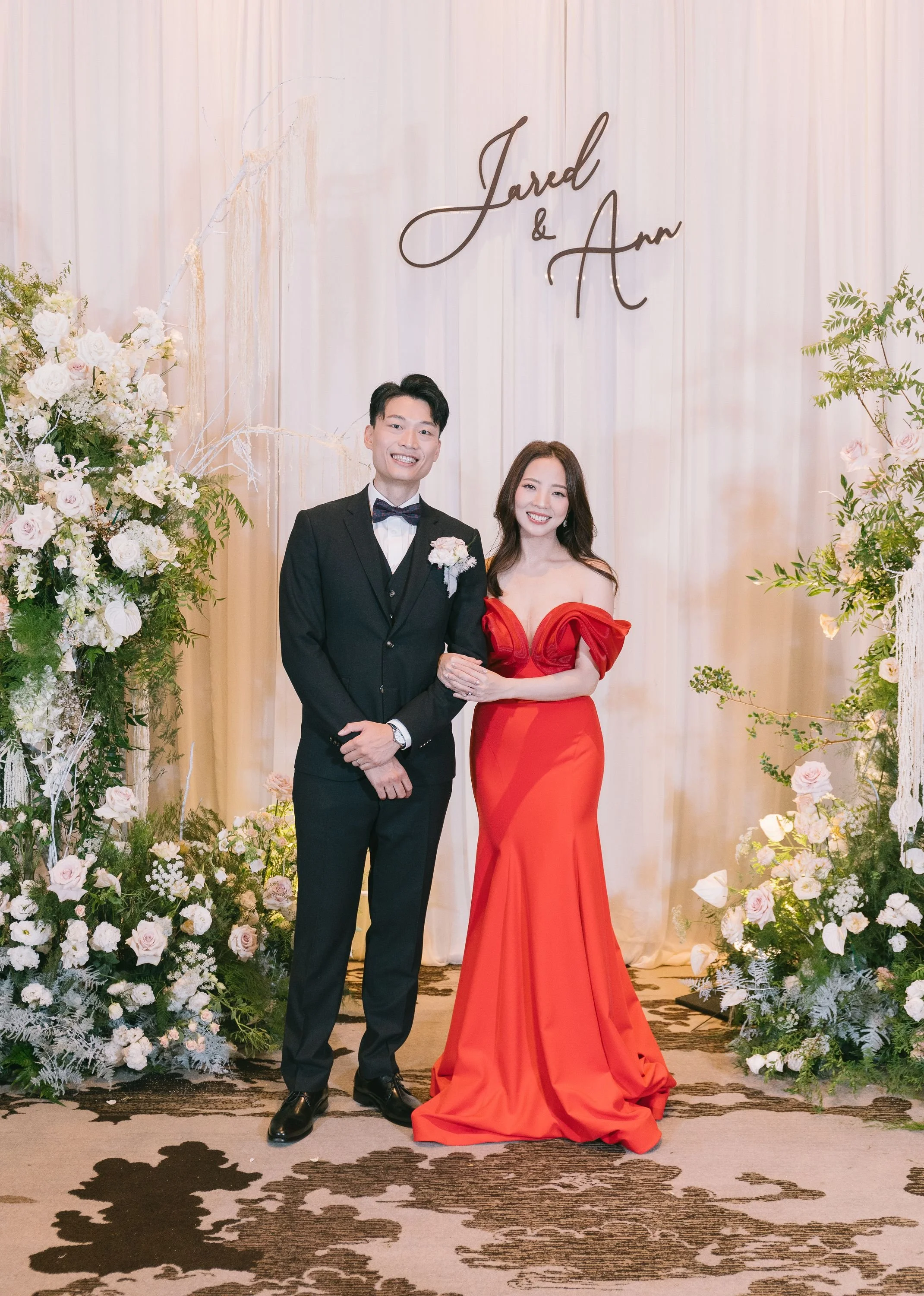 A smiling man in a black tuxedo with a bow tie and a woman in a red evening gown with off-the-shoulder sleeves standing together at a wedding celebration. Behind them is a decorated backdrop with white curtains, floral arrangements, and a sign with t