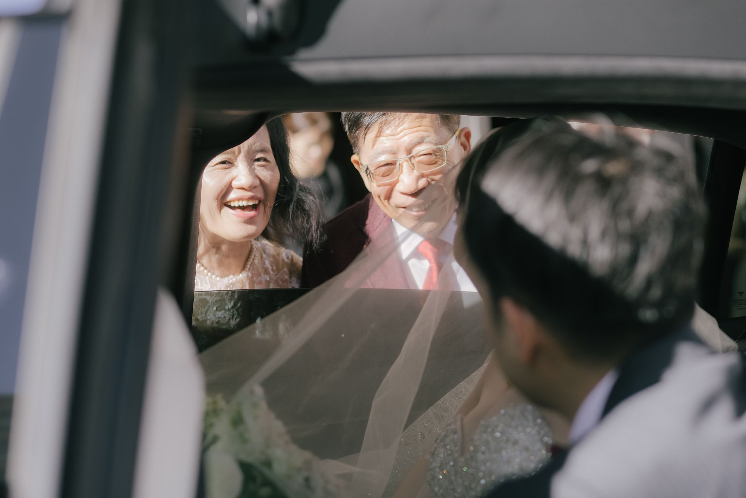 Smiling bride and groom looking at each other through a car window, with their wedding attire visible.