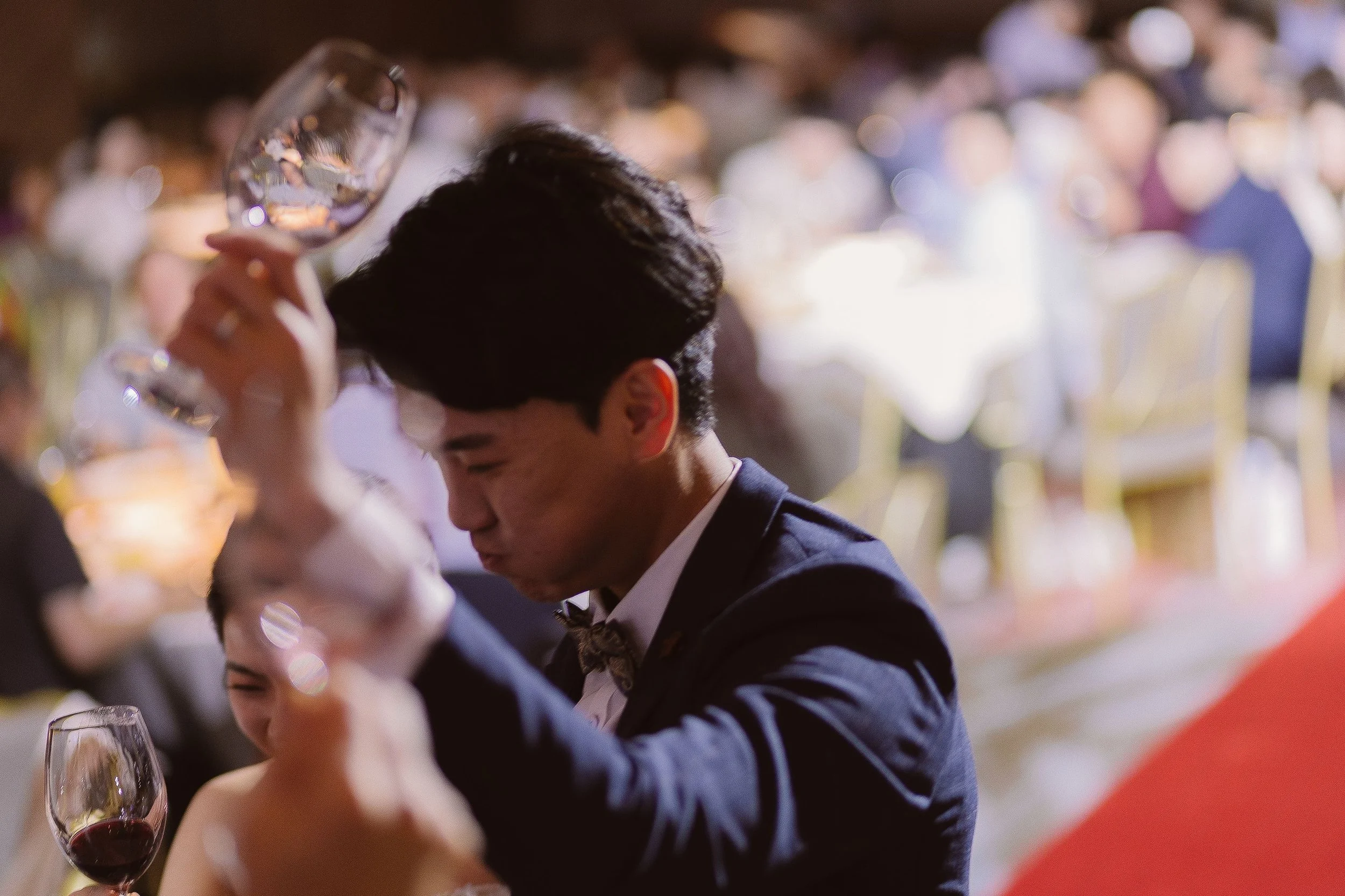 A man in a tuxedo sitting at a table with a glass of red wine. The background is blurred with people and lights, suggesting a formal event or celebration.