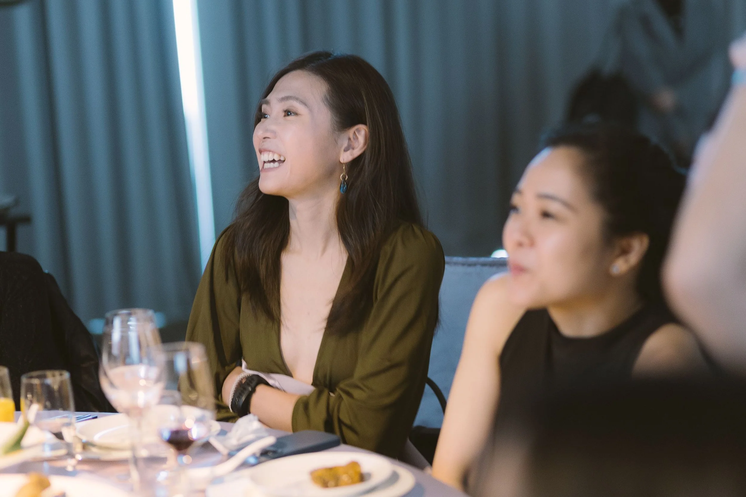 A woman with long dark hair laughing at a dinner party, seated at a table with wine glasses and food, in a room with blue curtains.