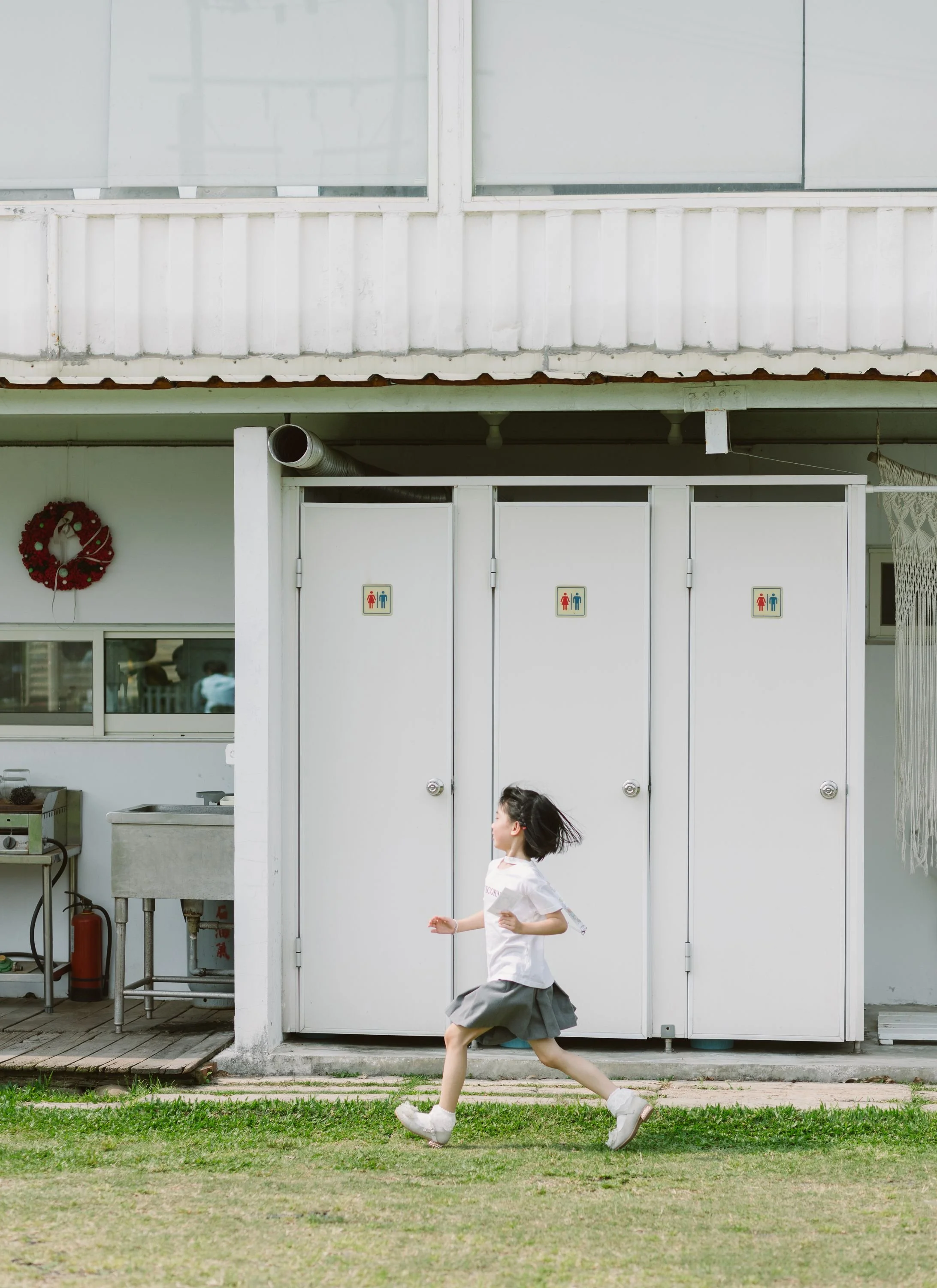 A young girl running on a grassy yard in front of a white building with a painted door. The girl is wearing a white shirt and a gray skirt, with white shoes, and has short dark hair.