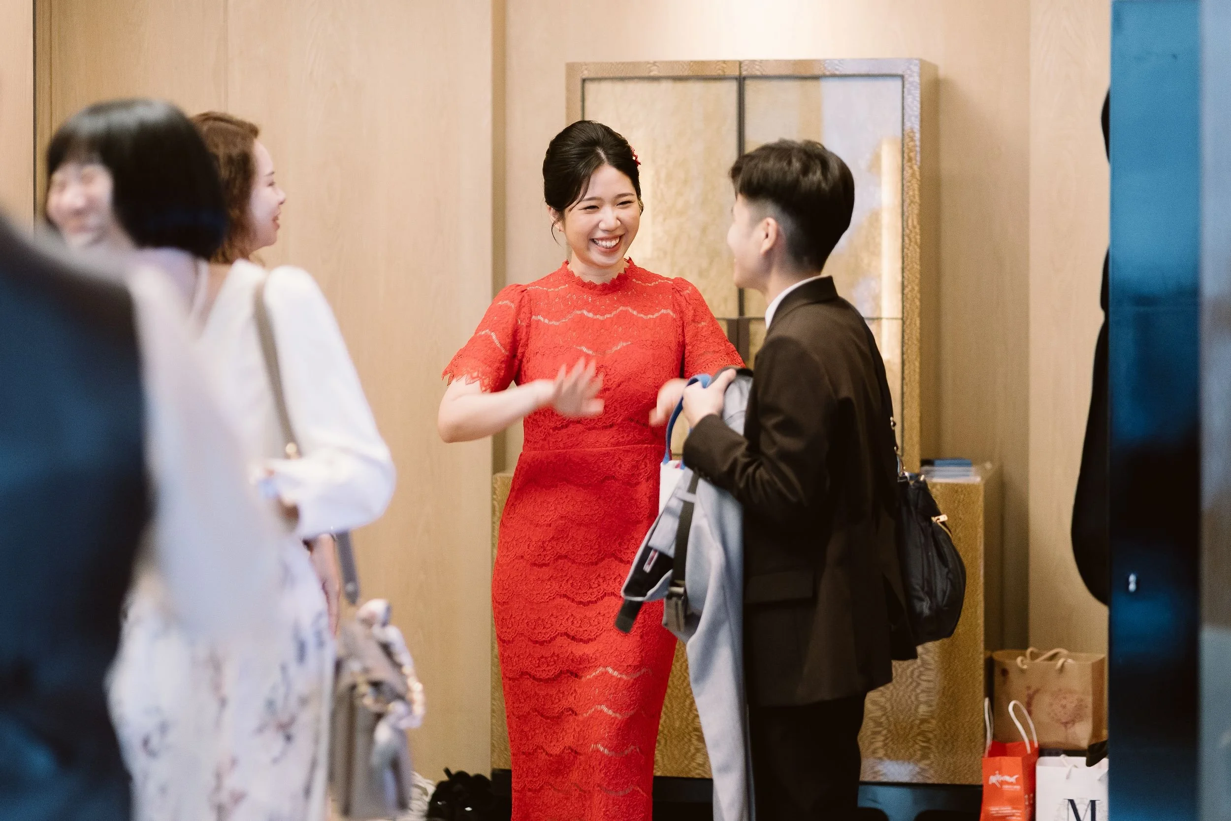 A woman in a red lace dress is smiling and talking to a young man carrying a backpack. Other women are visible in the background, engaged in conversation, in an indoor setting.
