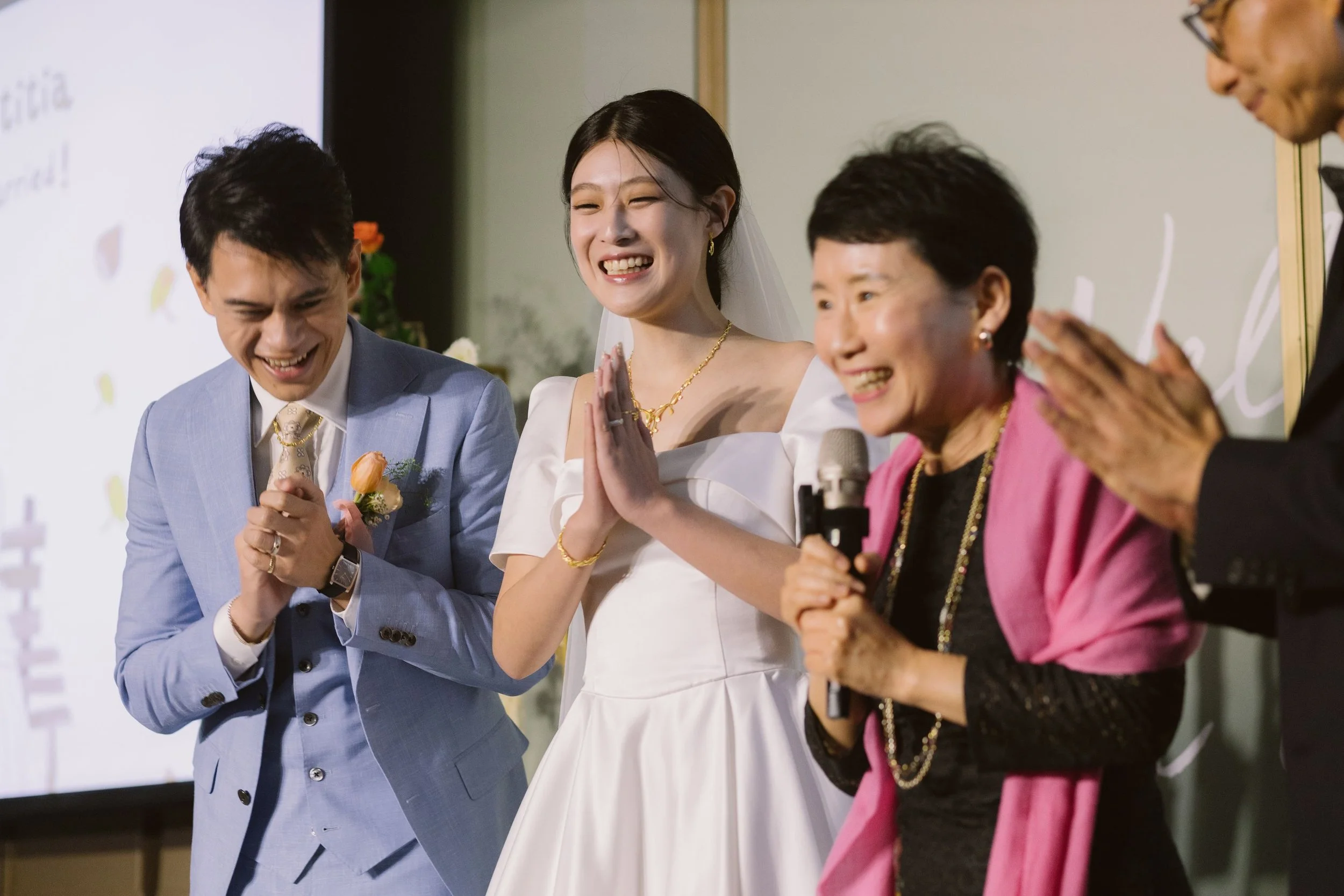 A group of five people, including a bride in a white dress, are smiling and praying with their hands pressed together during a wedding ceremony.