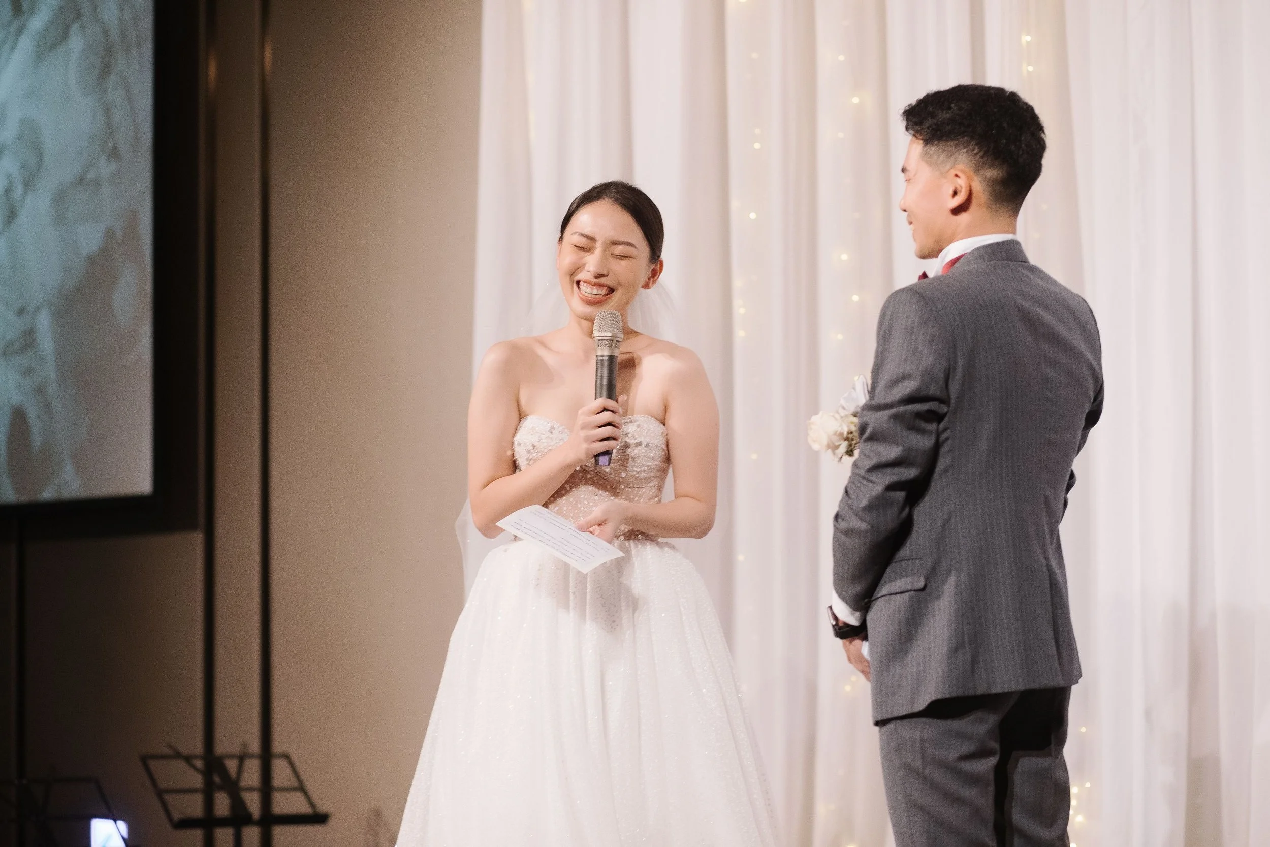 A bride with dark hair in a bun, wearing a strapless white wedding dress, holding a microphone and a piece of paper, smiling with eyes closed, standing next to a groom with short dark hair, wearing a gray suit with a white shirt and red bow tie, in f