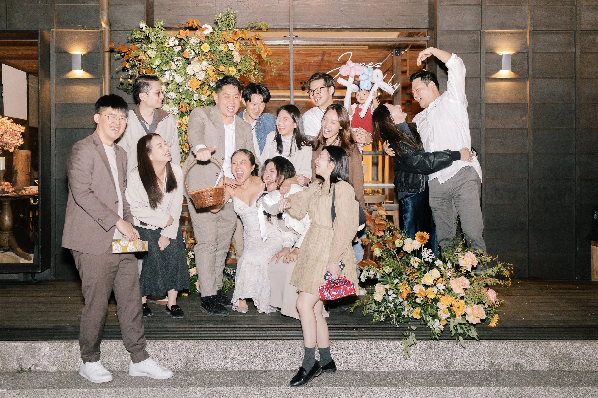 A group of people at a celebration, standing outdoors in front of a decorated floral backdrop. They are smiling, laughing, and interacting, with some holding balloons shaped like animals. There are flowers and greenery on the ground and in the backgr