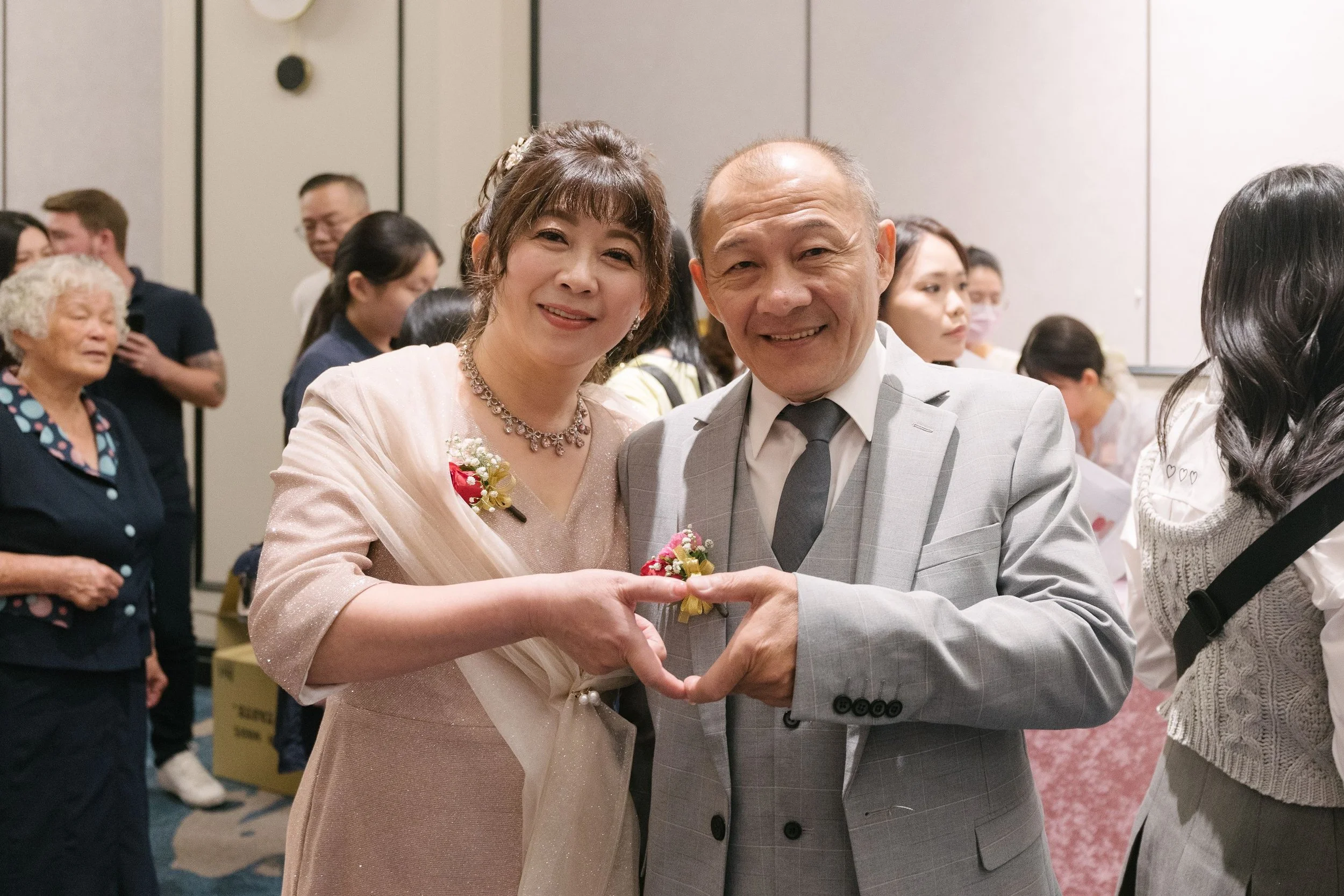 A man and woman are smiling and forming a heart shape with their hands at a social event.