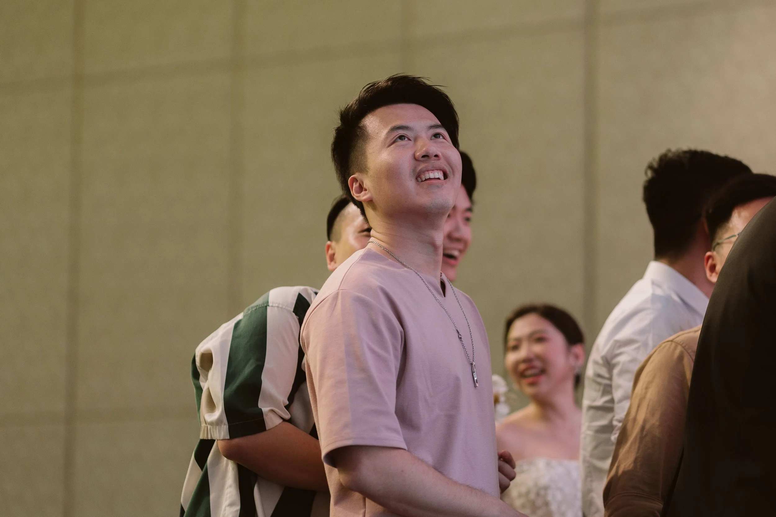 A young man with dark hair smiling and looking upwards at an event, surrounded by other people in a large room with beige walls.