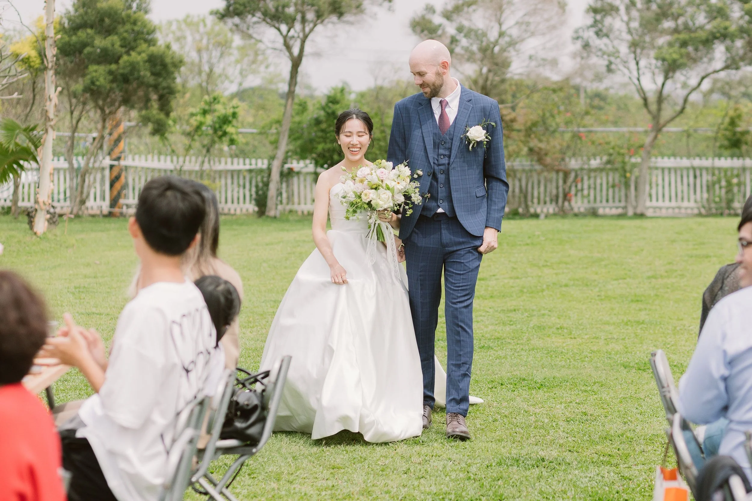 A wedding ceremony outdoors with a bride holding a bouquet, smiling, standing next to a groom in a blue checkered suit with a white flower boutonniere. Guests are seated on chairs, and trees and a white picket fence are in the background.