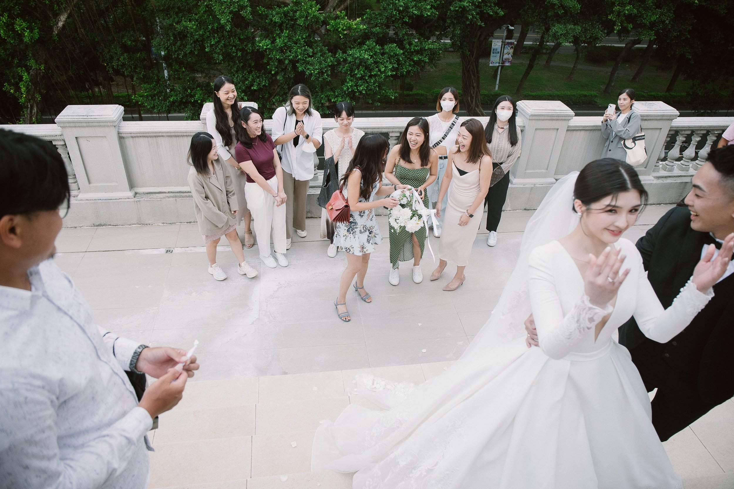 A group of people, including women in dresses and masks, celebrating a wedding with a bride and groom, on a balcony with a green park background.