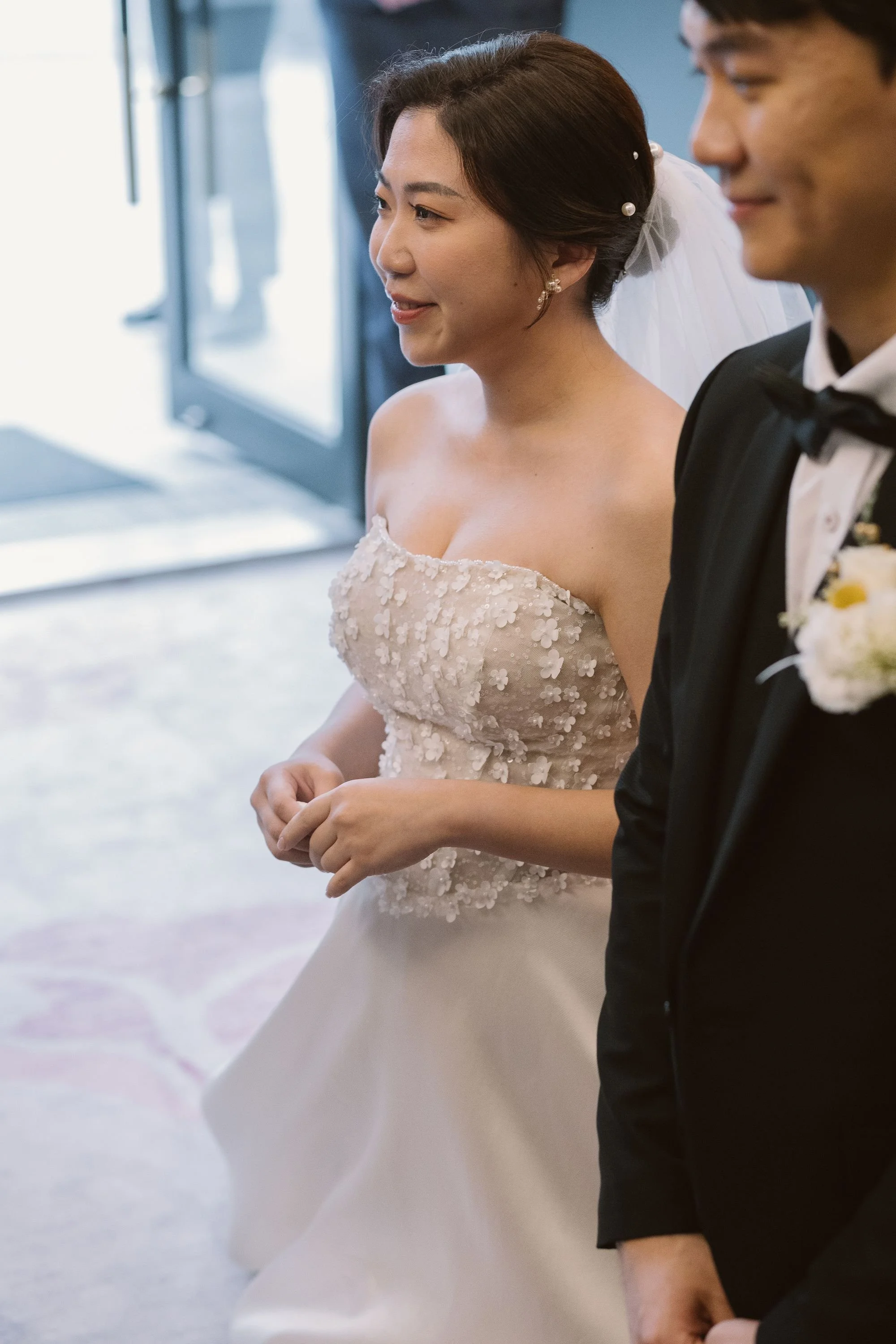 A bride and groom standing side by side during their wedding ceremony, both smiling softly, with a blurred background and natural light.