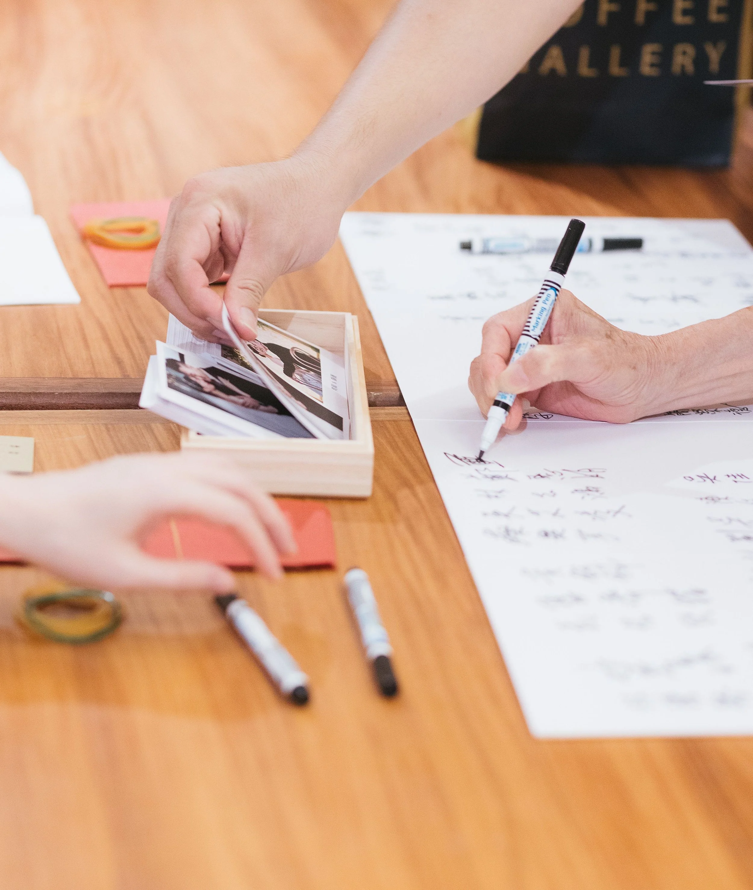 A person's hand is writing on white paper with a black marker. The paper contains handwritten notes and sketches. Other stationery items like whiteboard markers and post-it notes are on a wooden table nearby. A black gift bag with gold text is in the