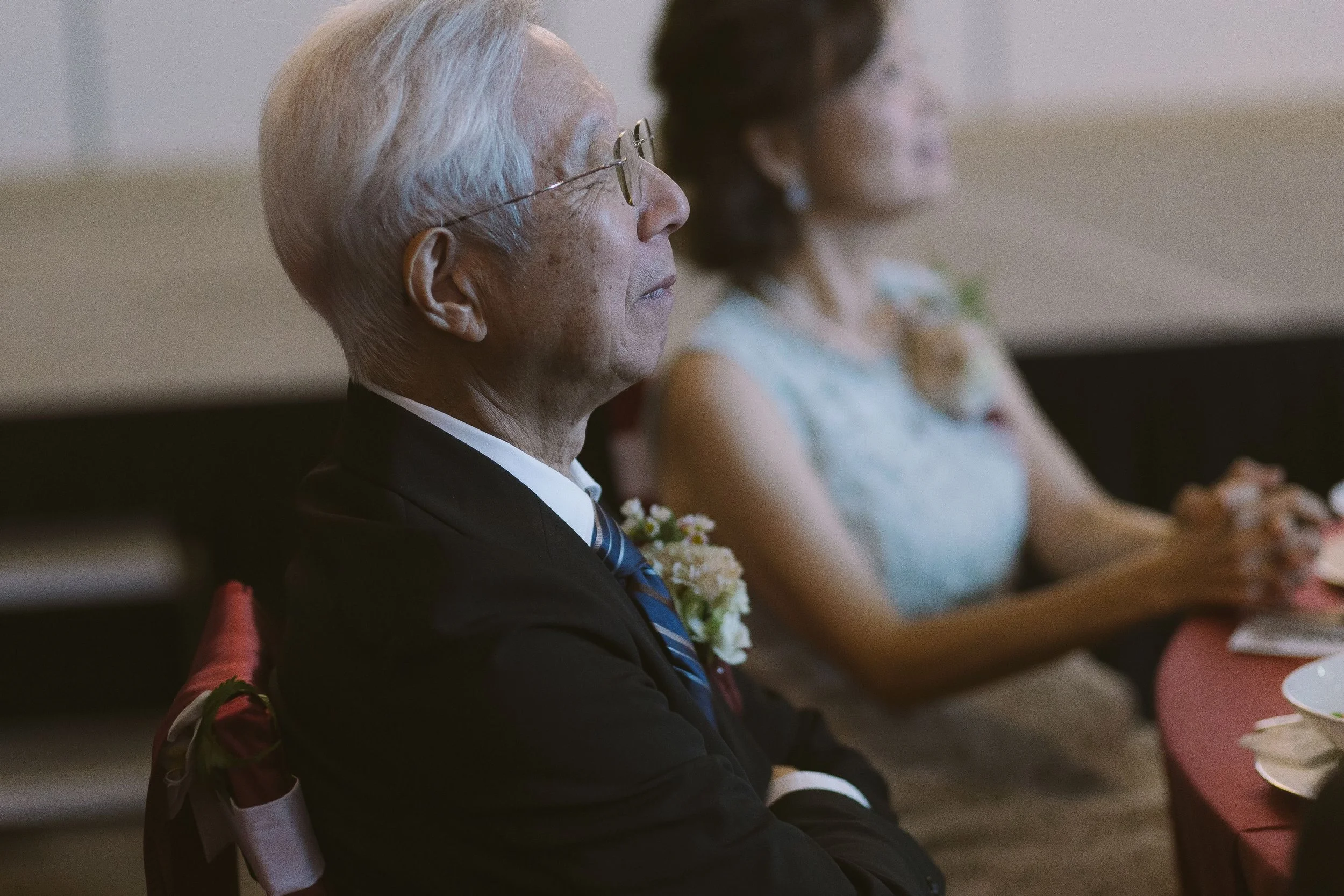 Side profile of an elderly man with gray hair and glasses, dressed in a black suit and blue tie, sitting at a formal event. Behind him, a woman in a light-colored dress is blurred in the background.