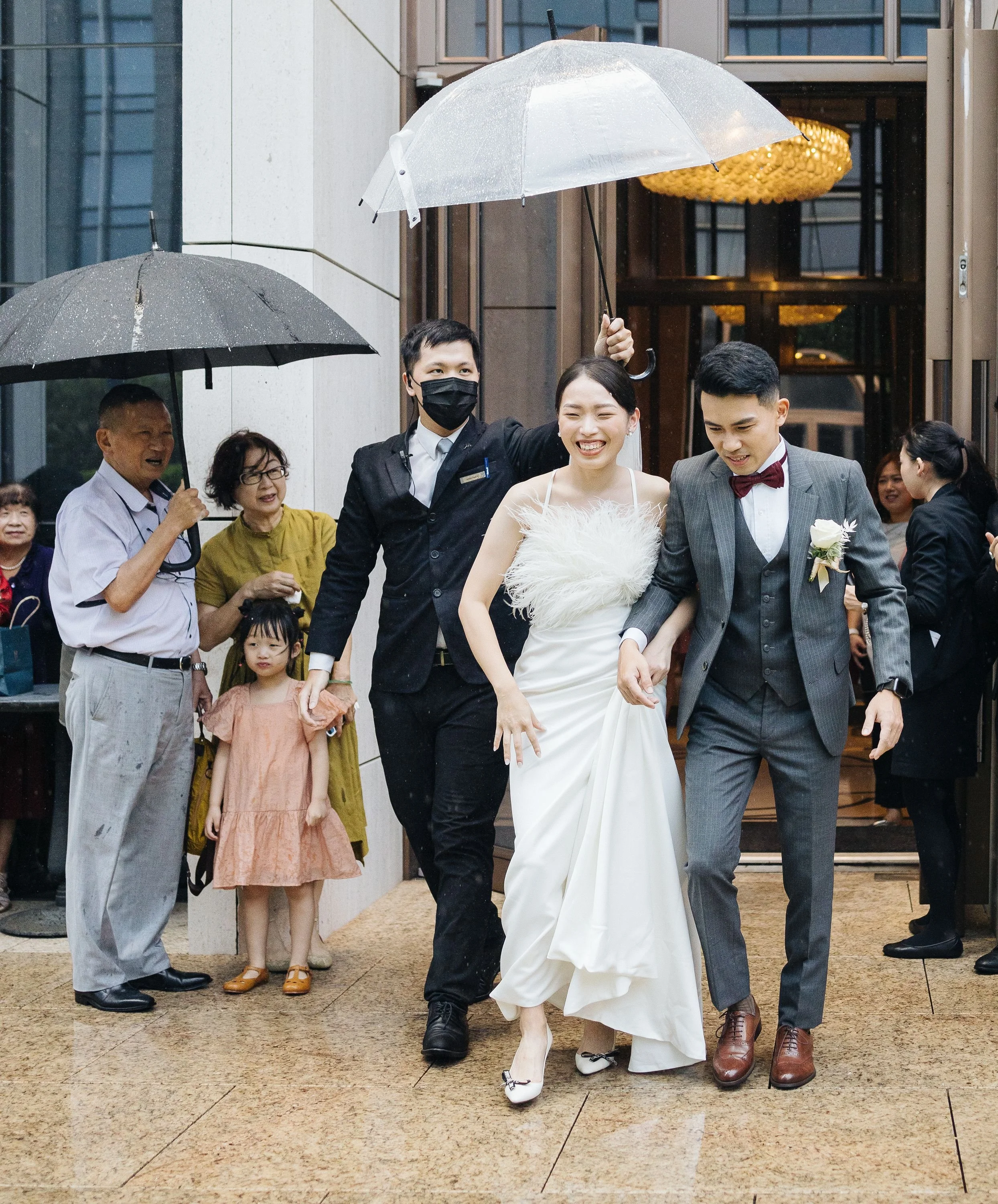 A bride and groom holding hands, walking outside during a rainy day, with people gathered around, some holding umbrellas. The bride is in a white dress with feather details, and the groom is in a gray suit with a bow tie.