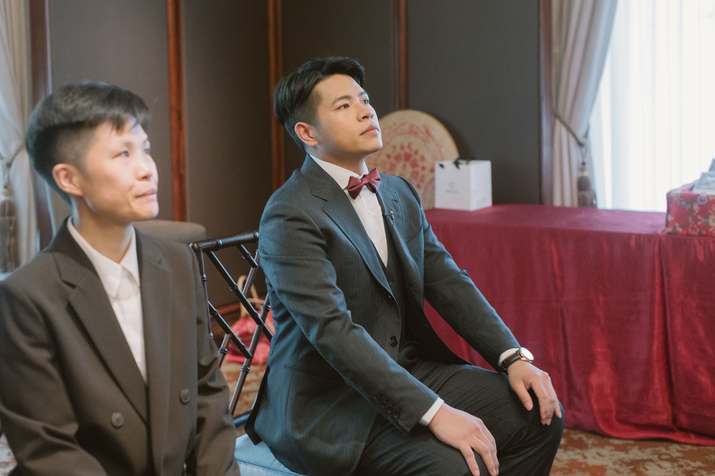 Two men dressed in business suits sitting on chairs indoors, paying attention to something, with a table and curtains in the background.