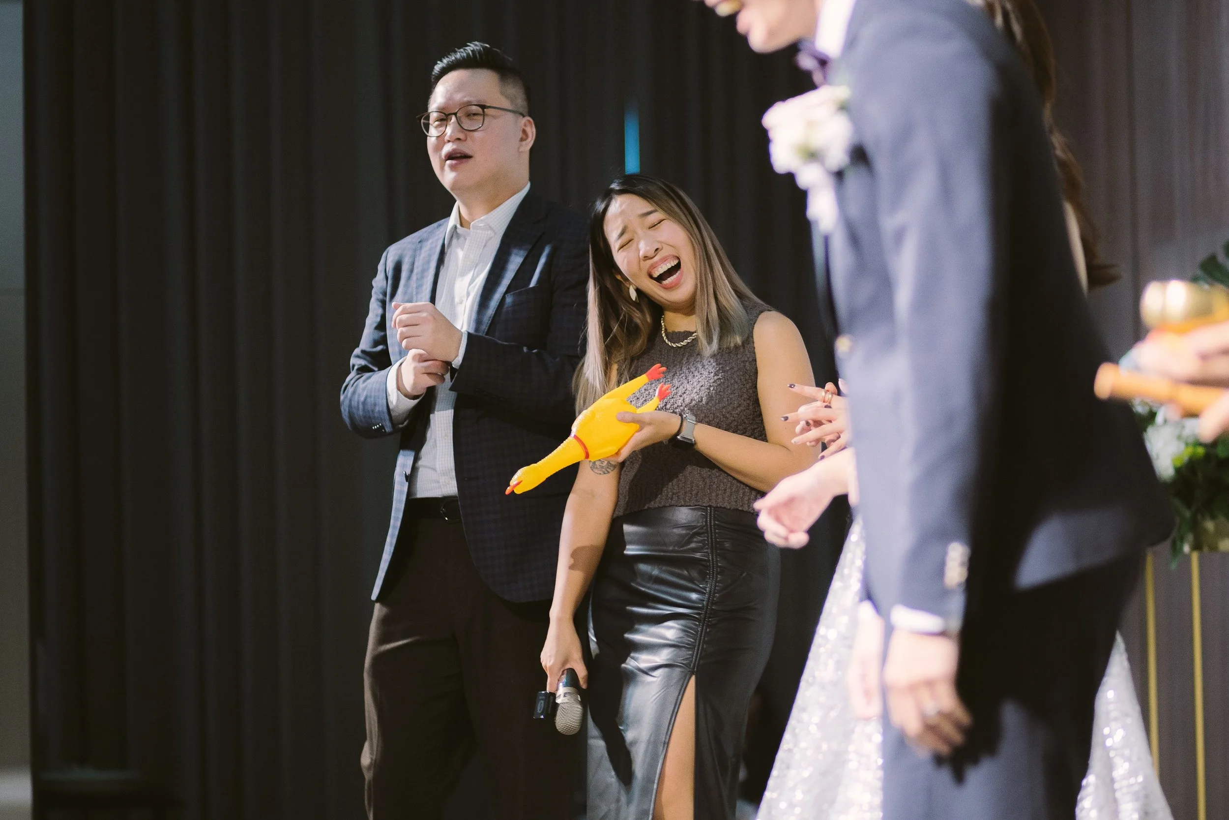 A group of people standing on a stage, some singing or speaking. A woman with long, dark hair is in the center, smiling and holding a yellow rubber chicken, wearing a leather skirt.