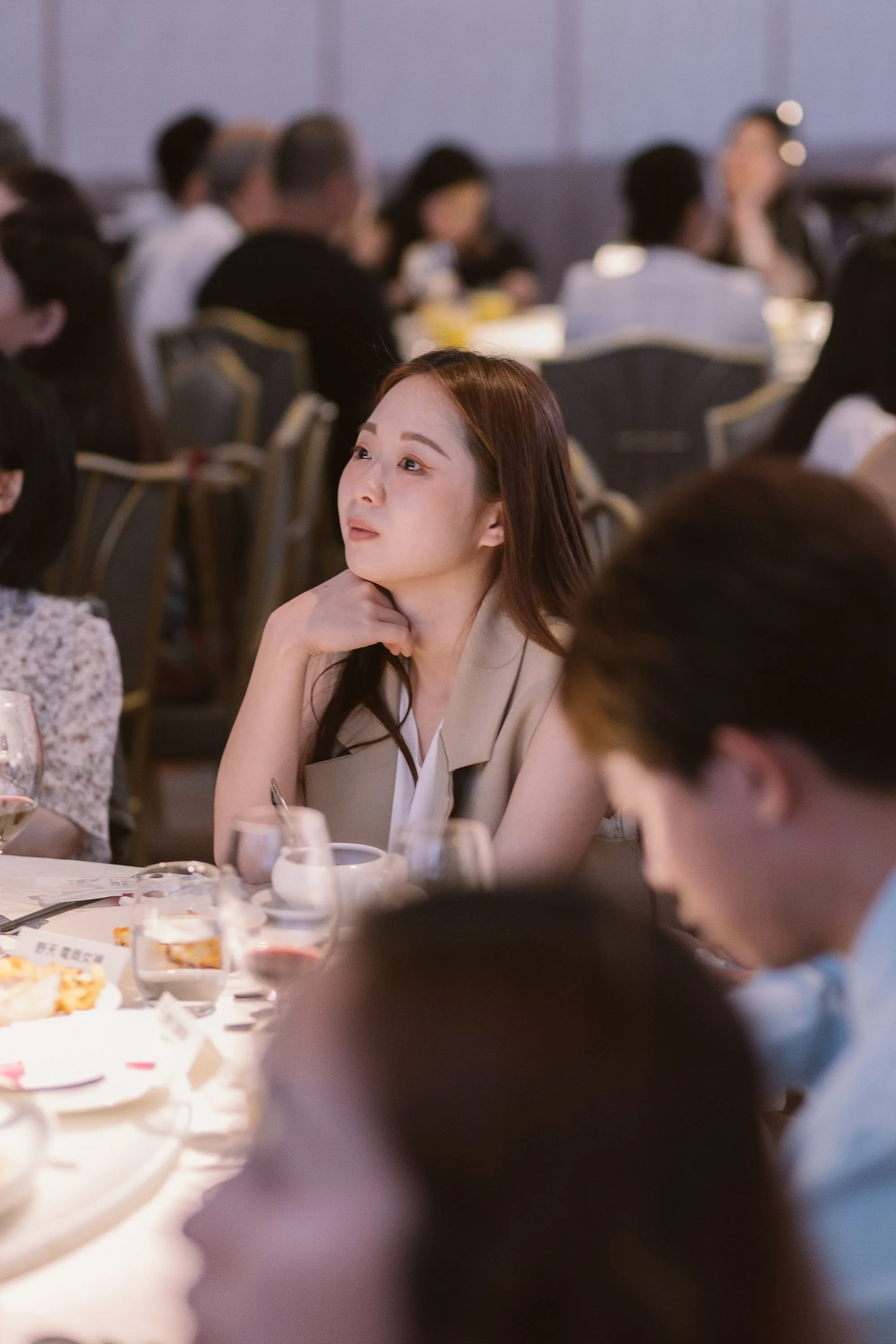A woman with long brown hair sitting at a banquet table during a formal event, resting her chin on her hand and looking thoughtfully to her right, surrounded by other guests.