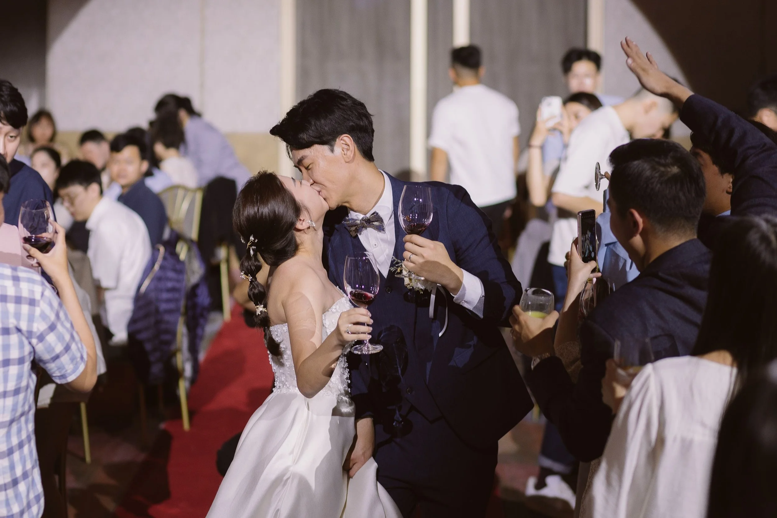 A wedding reception scene with a bride and groom sharing a kiss amid guests, some taking photos, in a decorated indoor venue.
