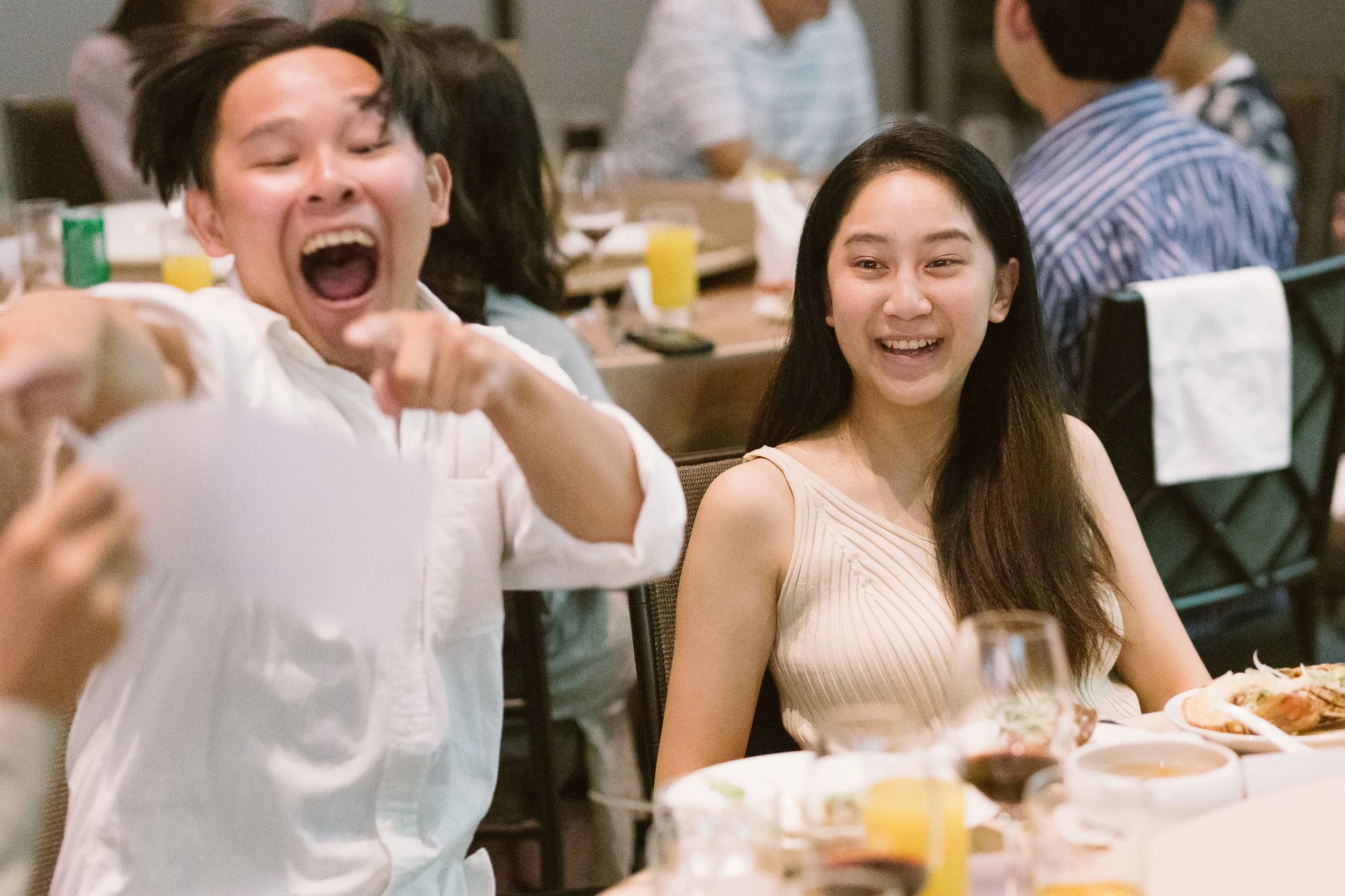 Happy young man with black hair laughing and pointing at the camera, sitting next to a smiling young woman with long black hair, in a restaurant with food and drinks on the table.