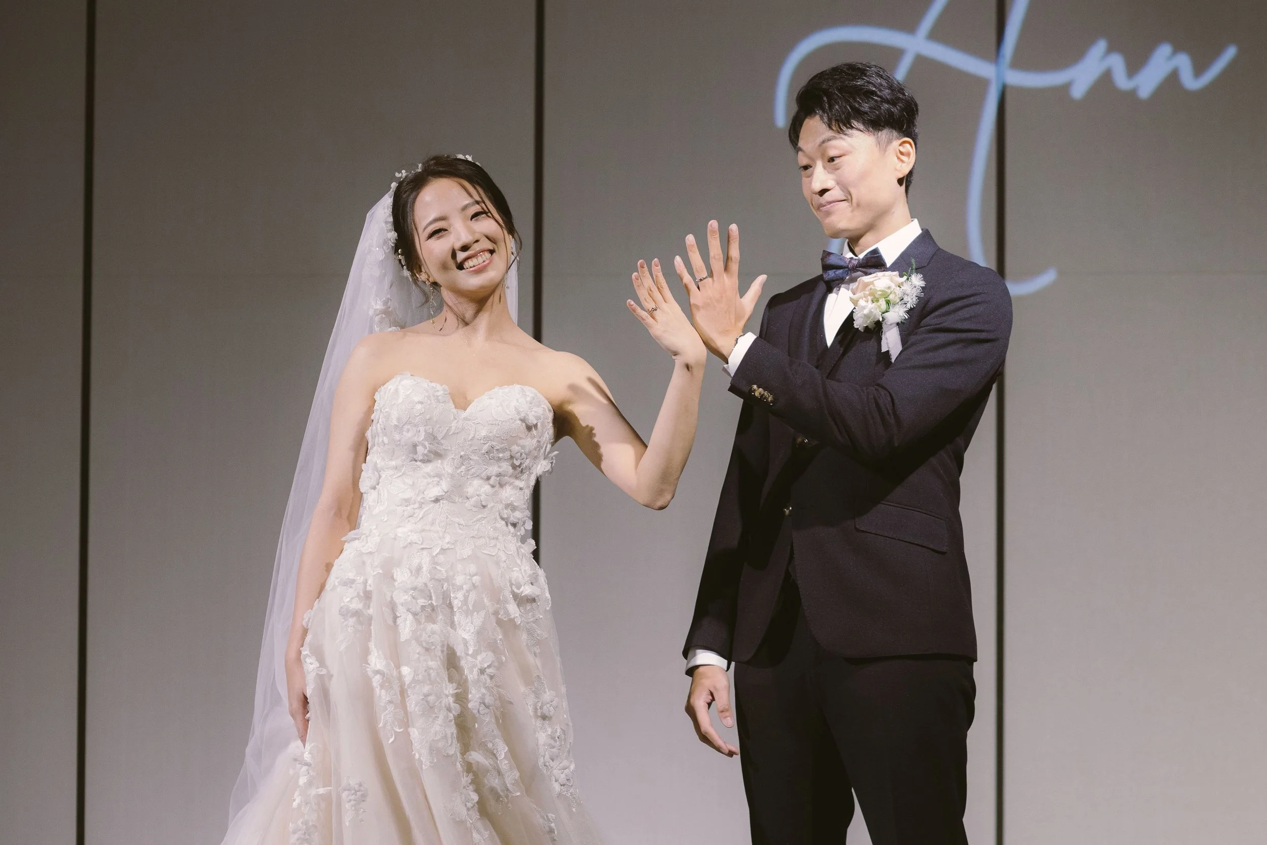 A couple dressed in wedding attire, smiling and showing their wedding rings, during their wedding ceremony or celebration.