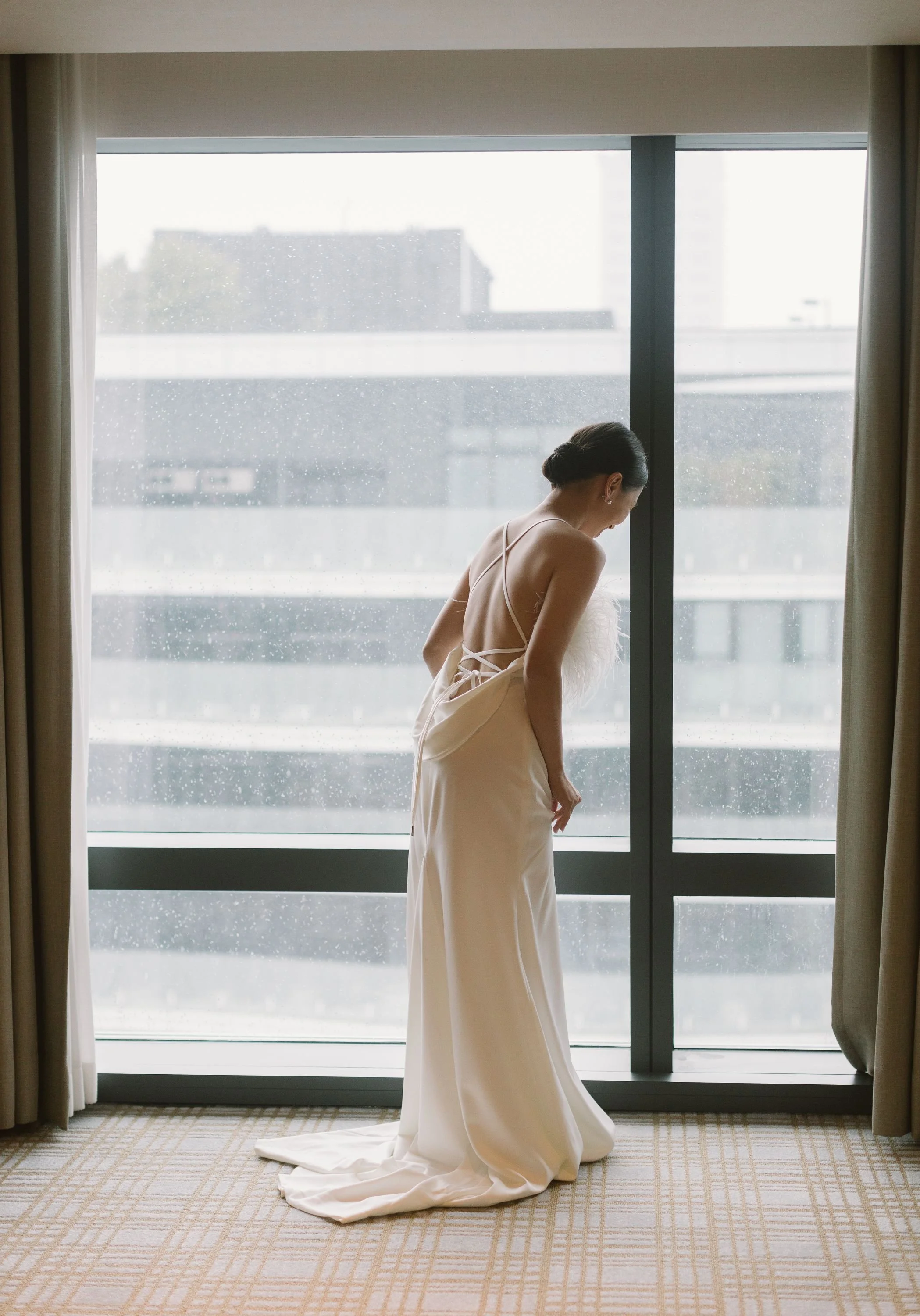A woman in a wedding dress standing by a large glass window on a rainy day, with city buildings in the background.