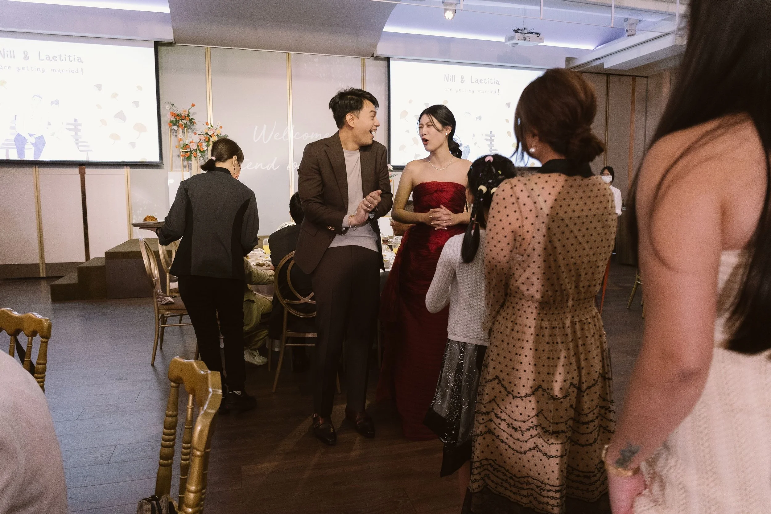 People gathered at a wedding reception, the bride and groom are in the center, smiling and chatting with guests. The bride wears a red dress, and the groom wears a brown suit. In the background, there are flower arrangements, a large welcome message 