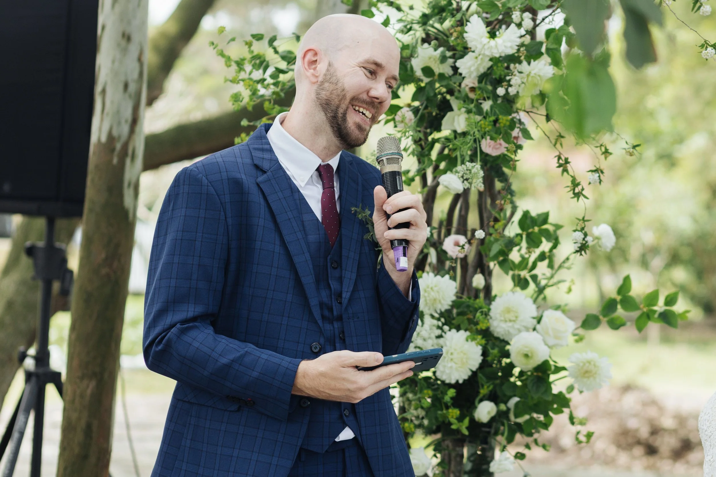 A man in a blue checkered suit holding a microphone and a phone, smiling at a wedding or outdoor event with a floral arch and greenery in the background.