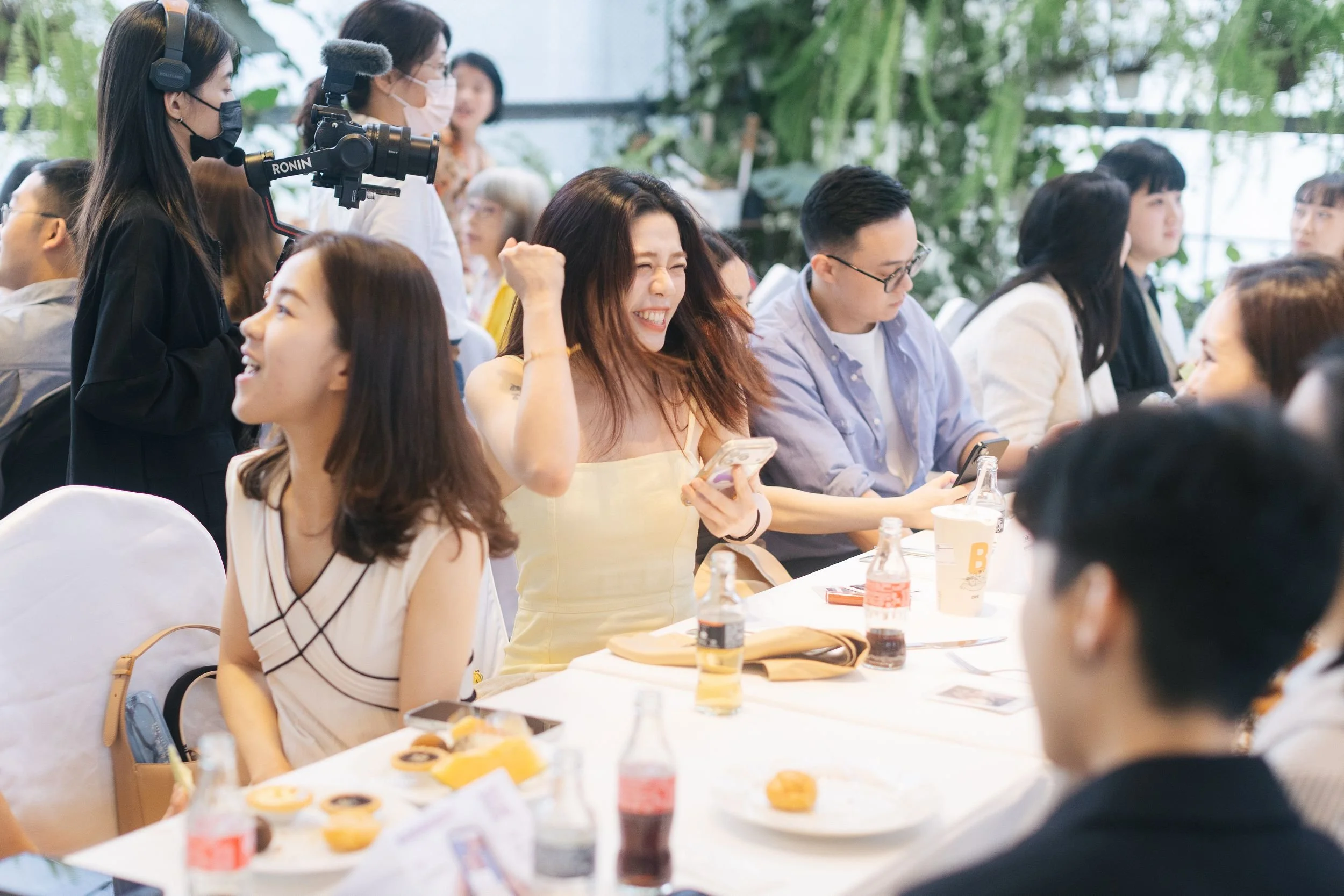 A group of diverse people attending an indoor event, with some eating and others engaging with their phones. A woman with a camera is recording the event, and the setting has lush green plants in the background.