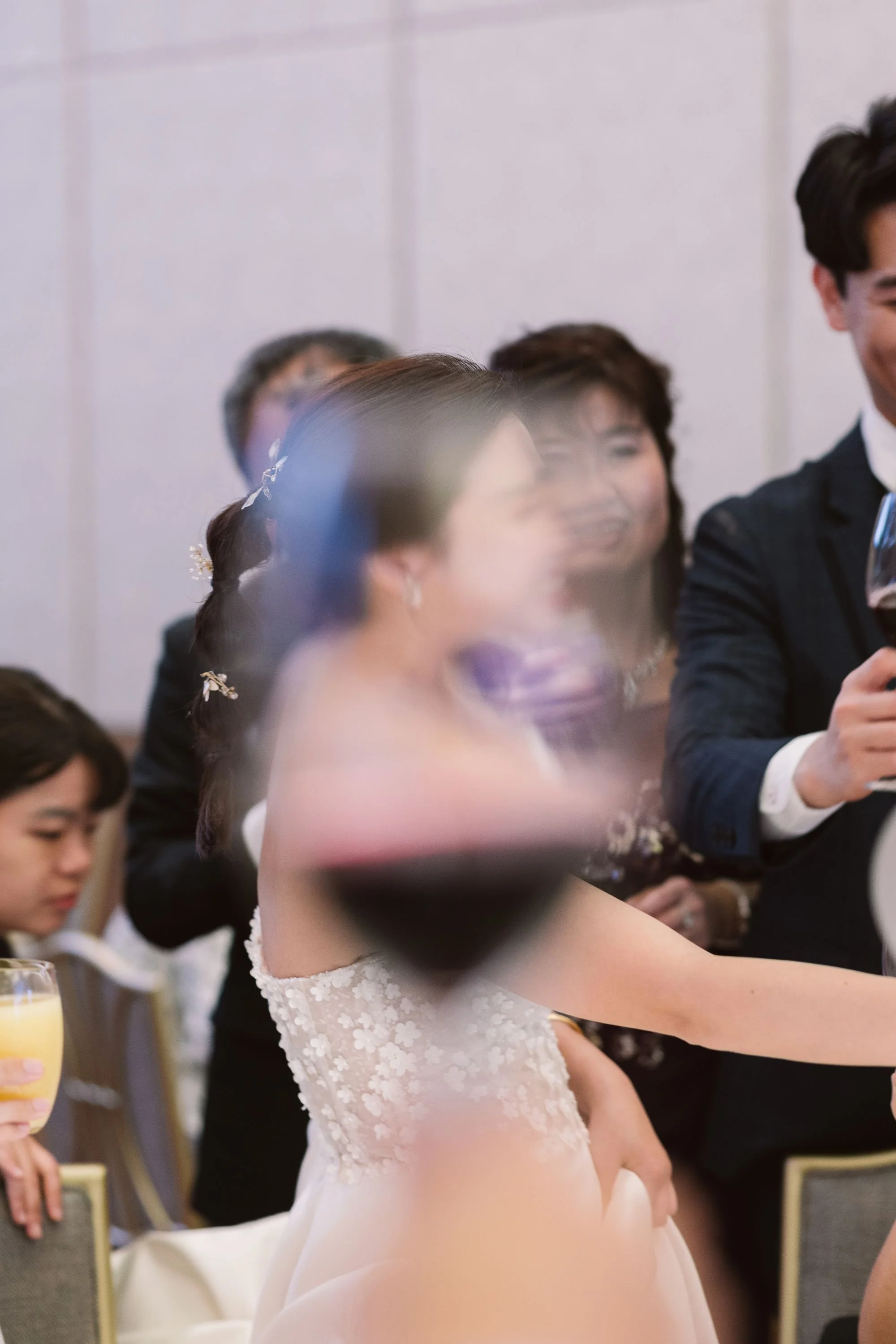 A bride in a white floral lace wedding dress at a wedding reception, smiling and holding a glass of orange juice, with guests seated at tables around her.