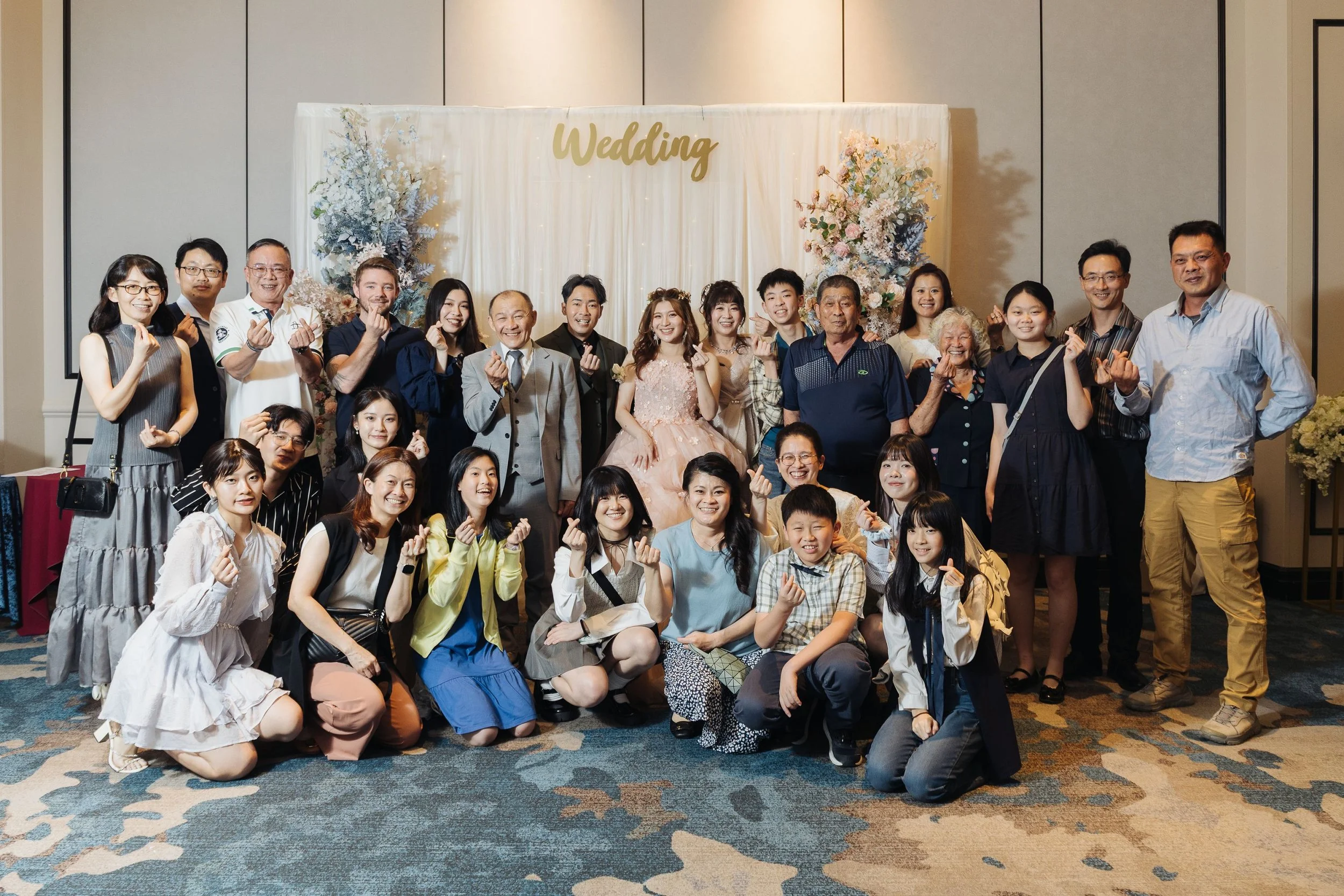 Group of people posing at a wedding celebration in front of a decorated backdrop with the word 'Wedding'.