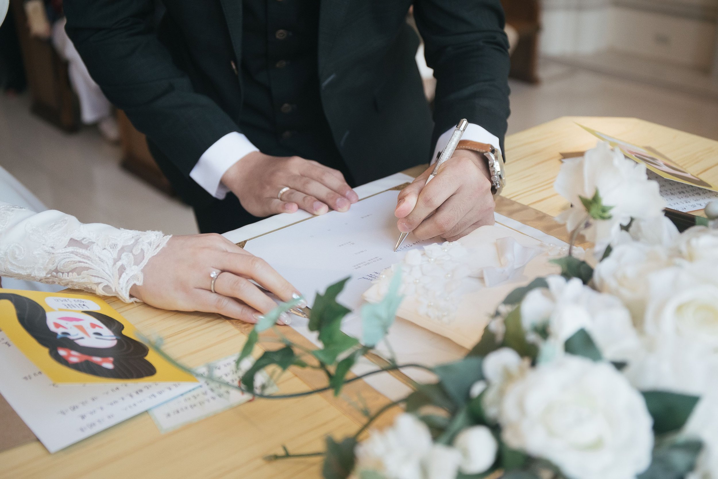A person in formal attire signing a document on a wooden table, with white flowers and colorful pamphlets nearby.