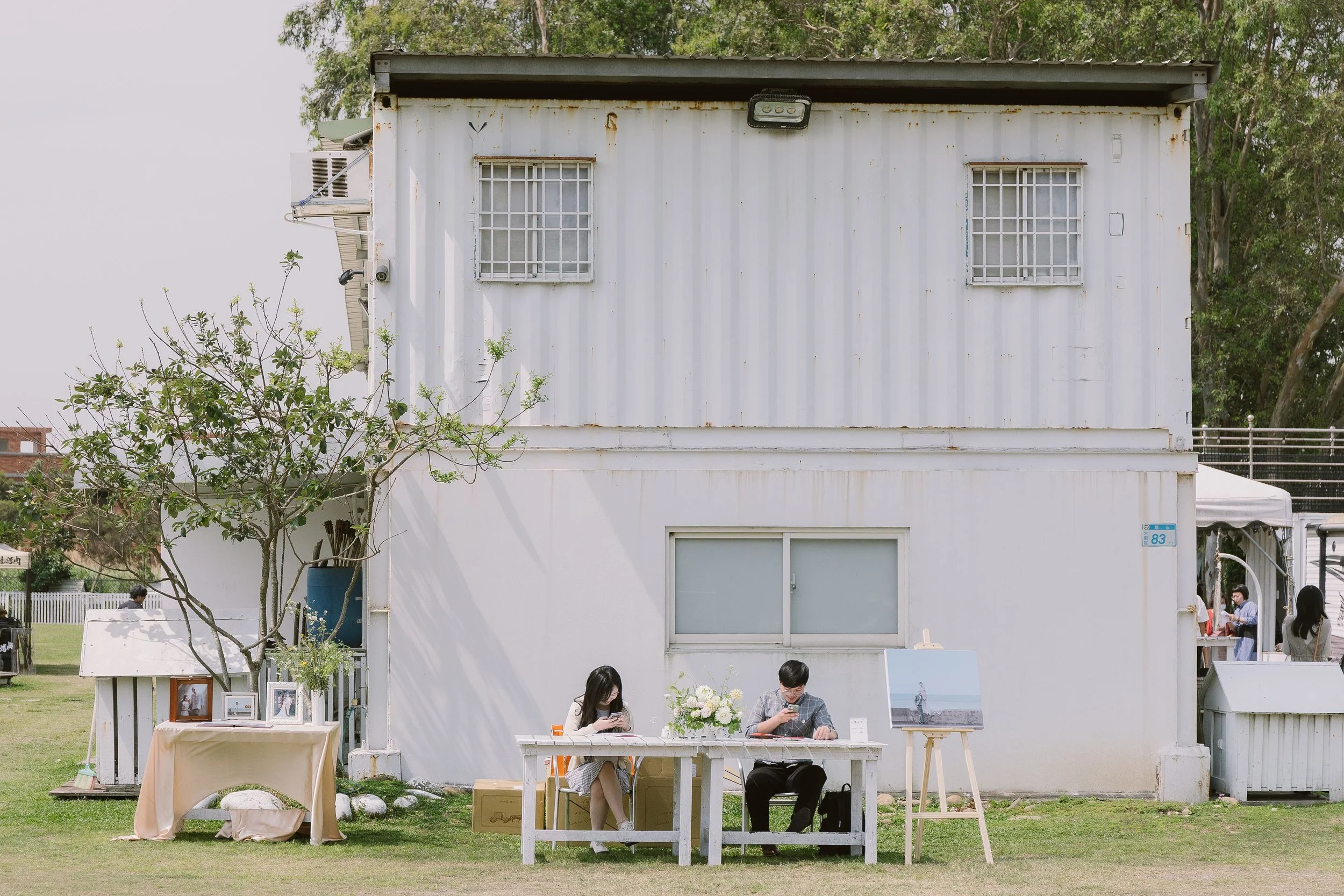A white building with two barred windows on the upper part and a large sliding window on the lower part, with trees and a fence in the background. In front, two people sit at a table with a flower arrangement, and there are other people and objects a