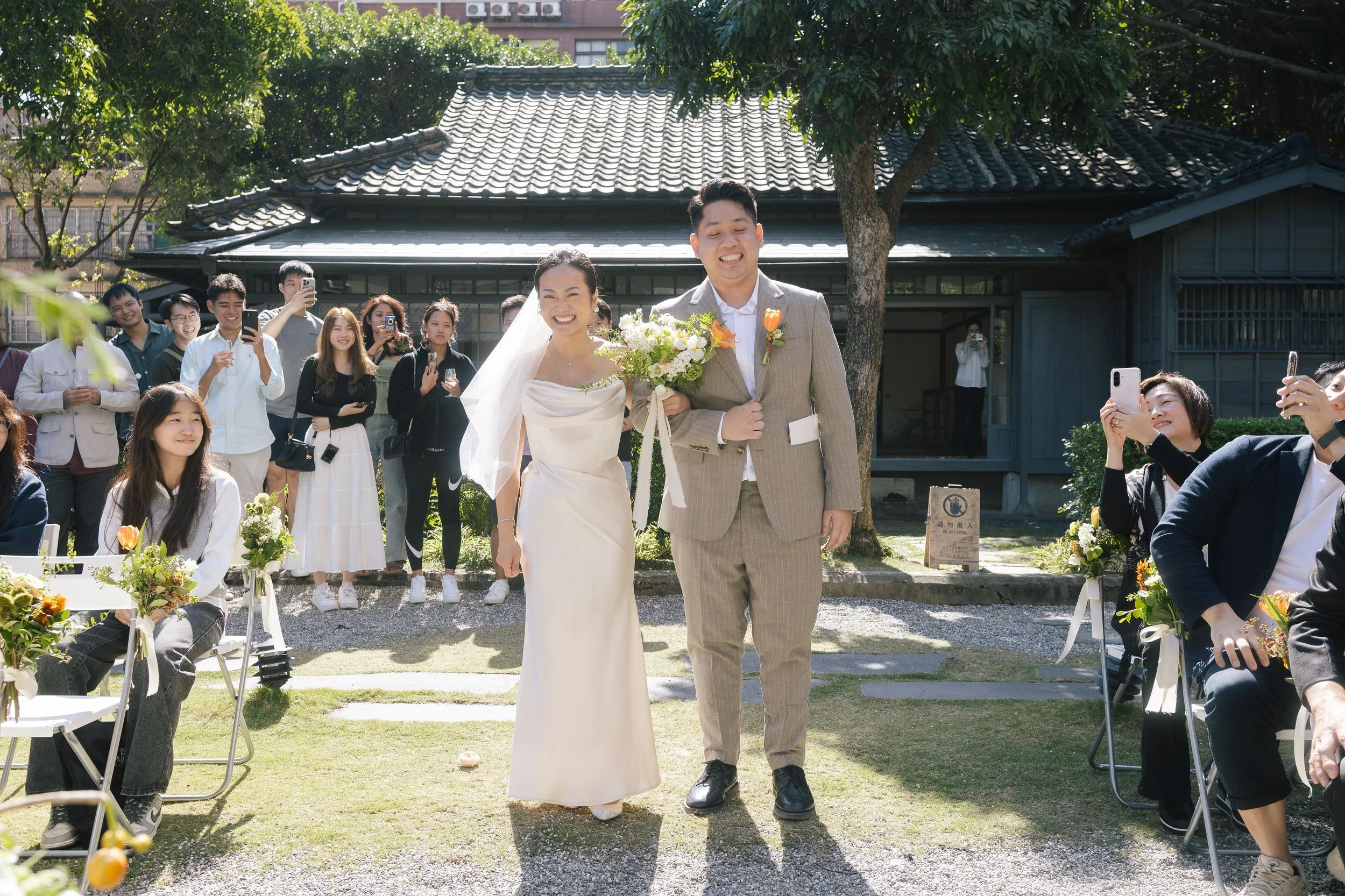 A bride and groom standing together outdoors at a wedding, smiling, with guests taking photos around them. The bride wears a white dress with a veil, and the groom wears a beige suit. The background features a traditional building with a tiled roof a