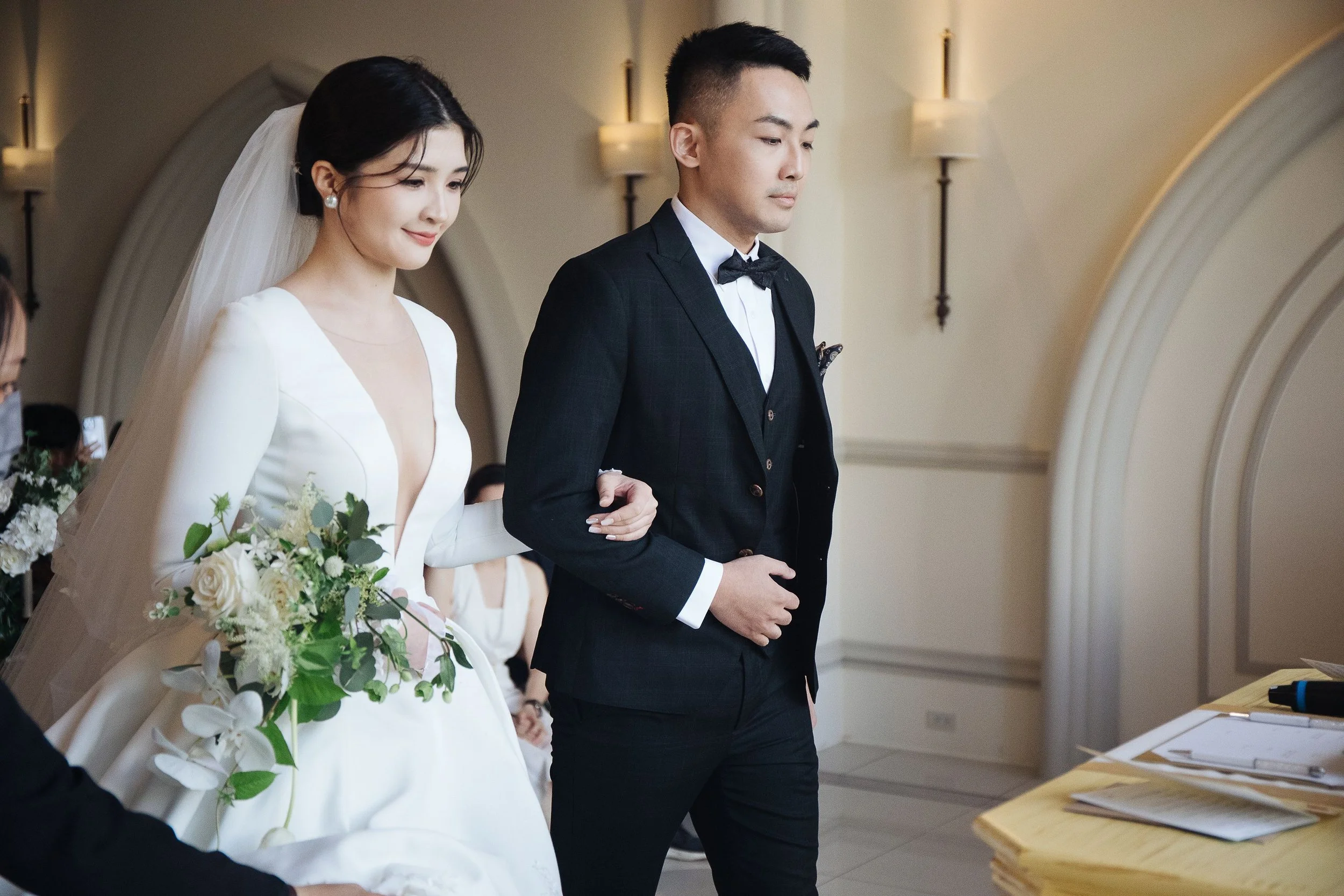 A bride and groom standing side by side during their wedding ceremony. The bride has dark hair and is wearing a white wedding dress with a veil, holding a bouquet of white and green flowers. The groom is dressed in a black suit with a white shirt and
