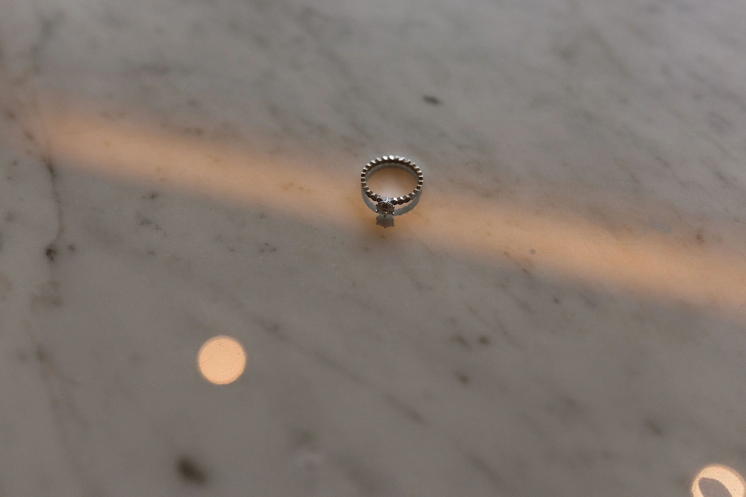 A silver ring with a central gemstone on a white marble surface with subtle veining, illuminated by soft lighting.