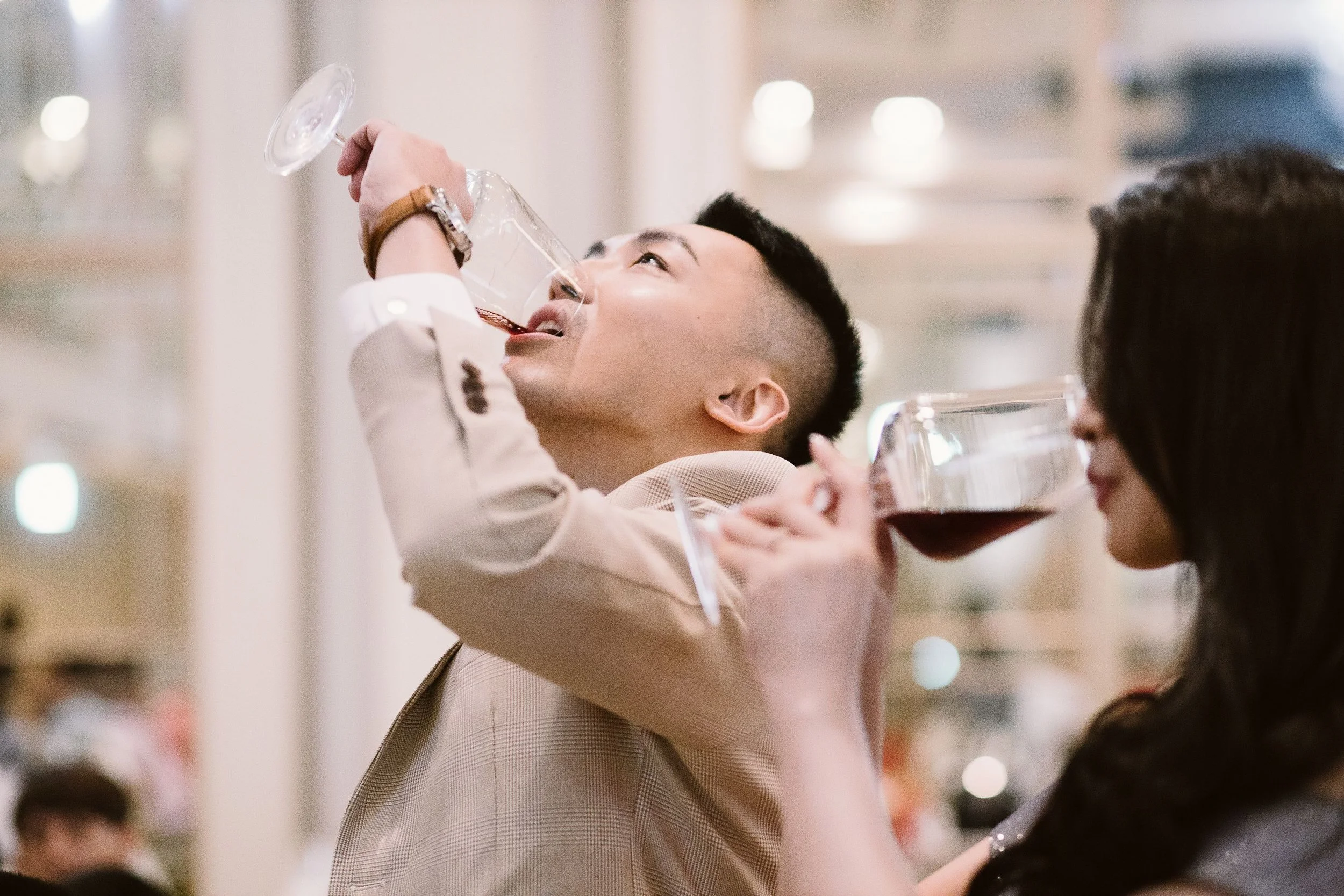 A young man in a light-colored blazer is drinking red wine from a glass at a social gathering, with other people visible around him.