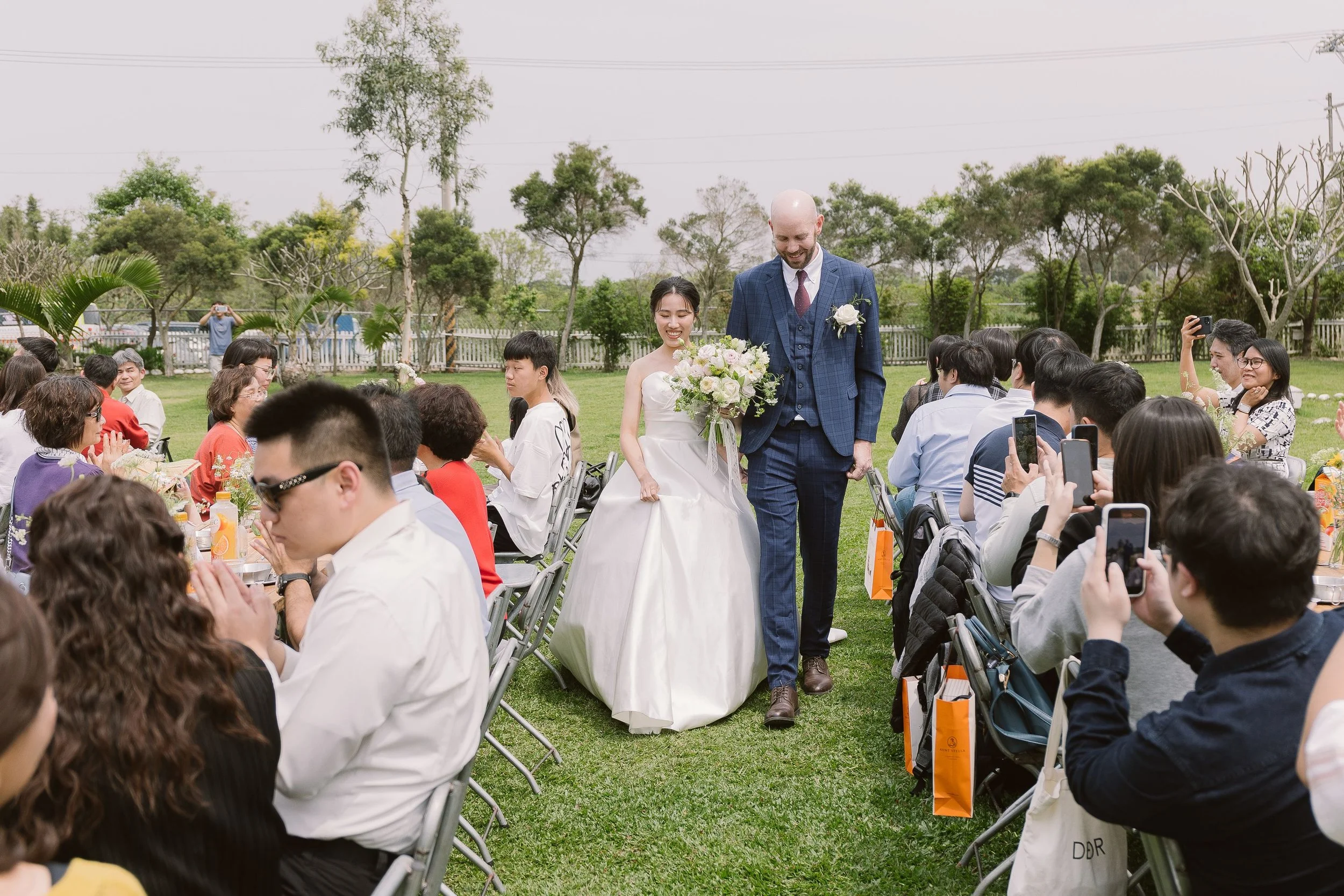 A wedding ceremony outdoors with a bride and groom walking down the aisle, surrounded by seated guests on a grassy area with trees in the background.