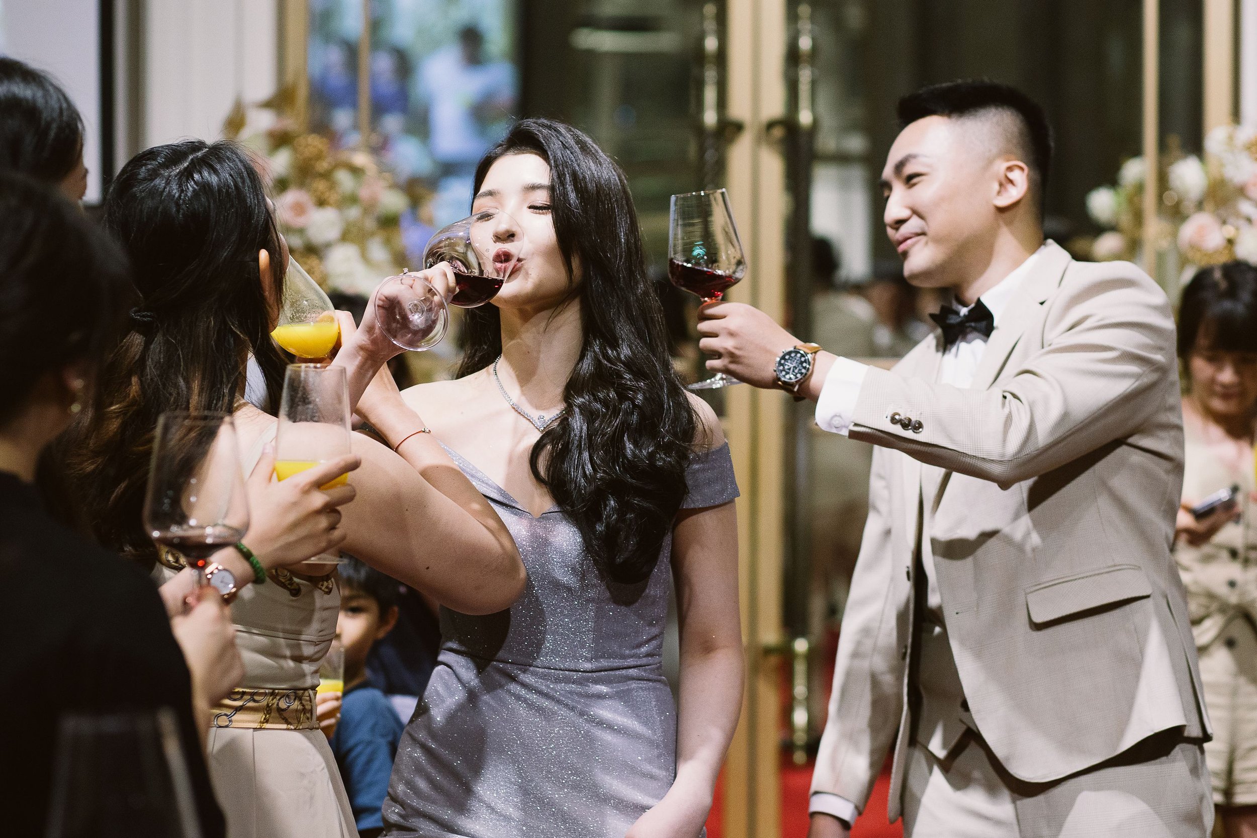 A woman in a silver dress is being tapped for wine by another woman at a social gathering, with a man in a white tuxedo raising a wine glass nearby. The scene is lively with other guests in the background.