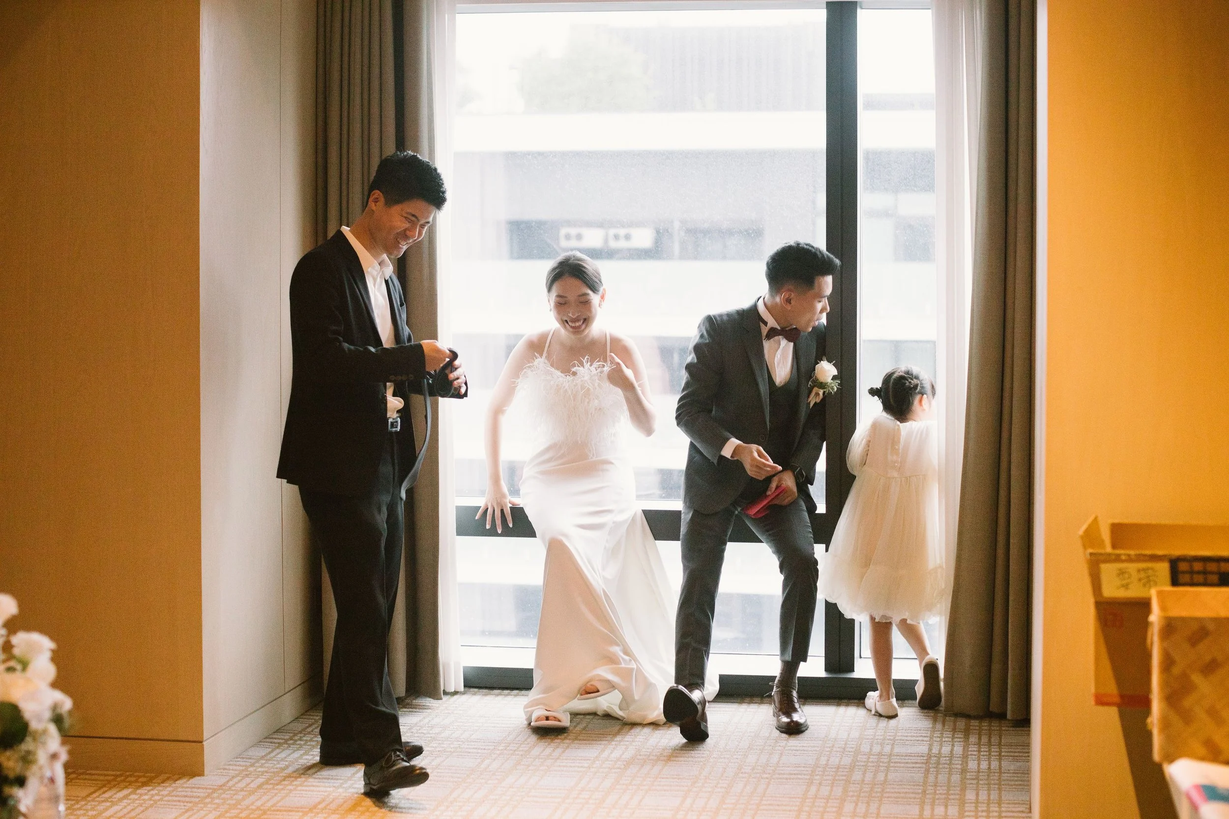 A group of four people dressed in formal attire, including a bride in a white gown, two men in suits, and a little girl in a white dress, are standing near an open door with a bright background outside. They appear to be smiling and enjoying a moment