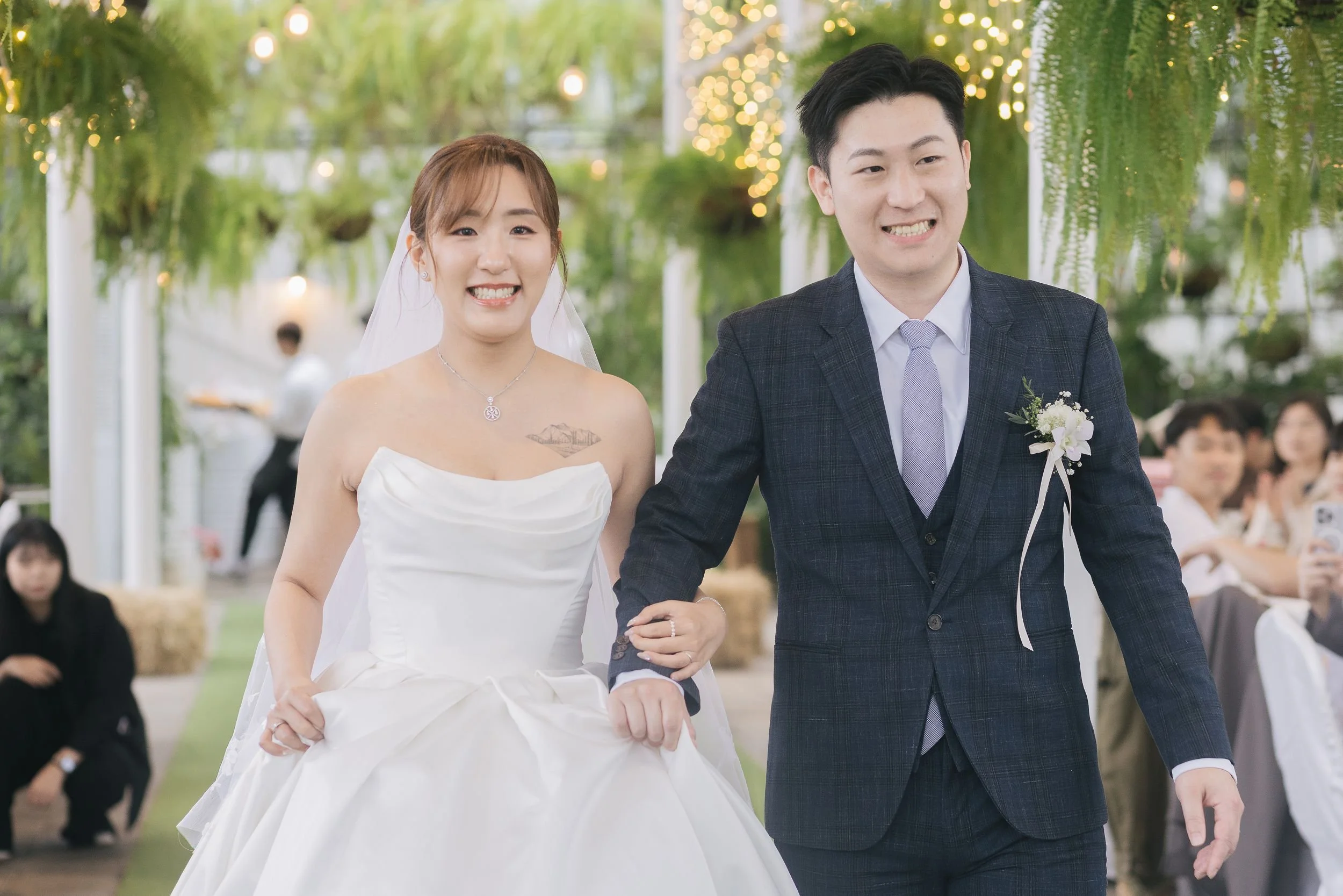 A smiling bride and groom walking arm in arm at their wedding reception, decorated with hanging greenery and string lights, with guests seated in the background.