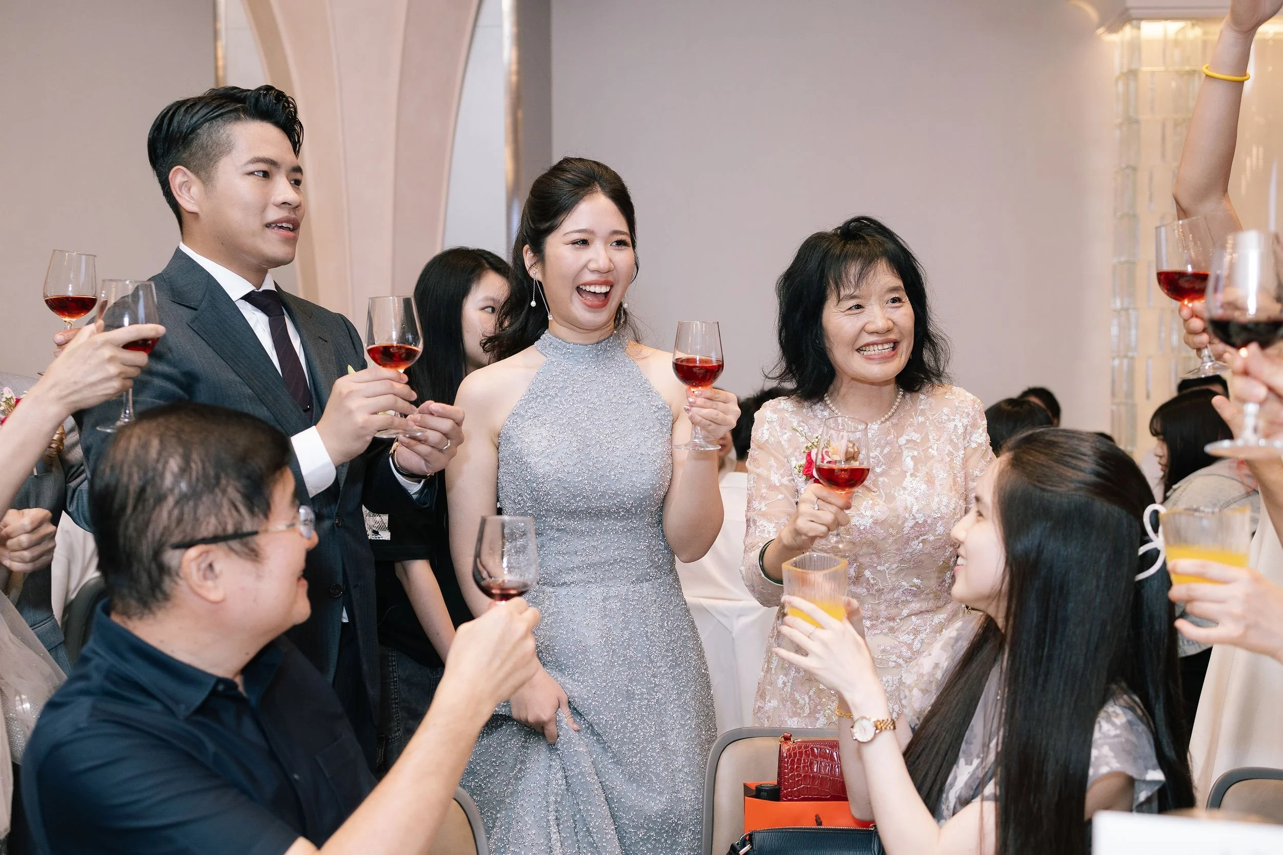 A group of people celebrating at a social gathering, holding glasses of red and orange beverages, smiling, and engaging with each other. The setting appears to be indoors at a festive event.