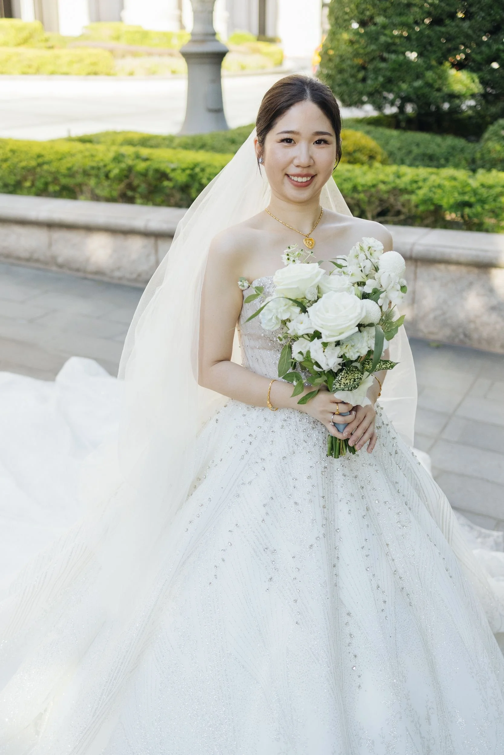 A smiling bride with dark hair in a wedding dress holding a white bouquet, standing outdoors near a stone wall and greenery.