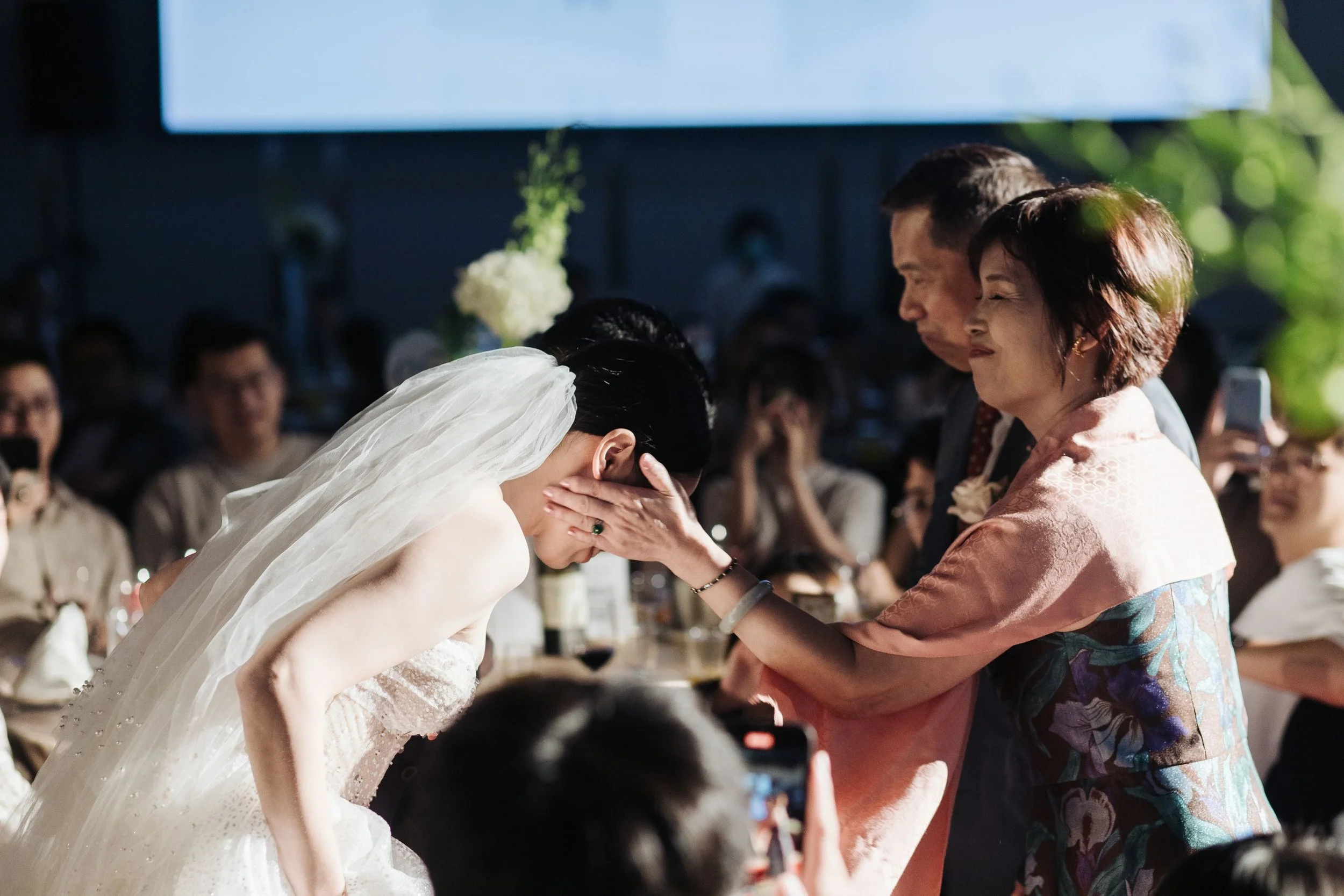 A wedding ceremony where a bride with a white veil and dress is kissing an elder woman, possibly her mother, who is wearing a traditional dress and holding the bride's face with both hands. Many guests are visible in the background, taking photos and