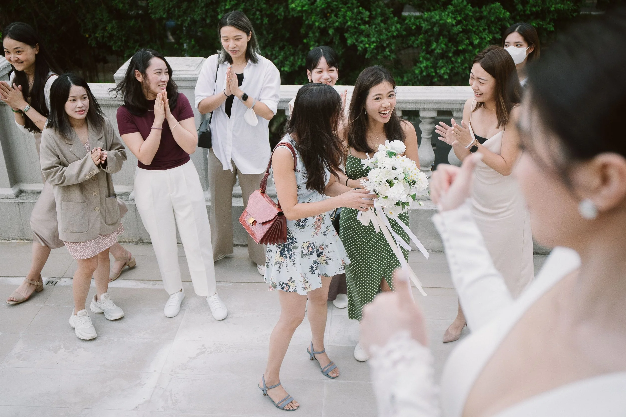 A group of women gather outdoors, some smiling and clapping, others with hands in prayer position, with greenery and a stone railing in the background.