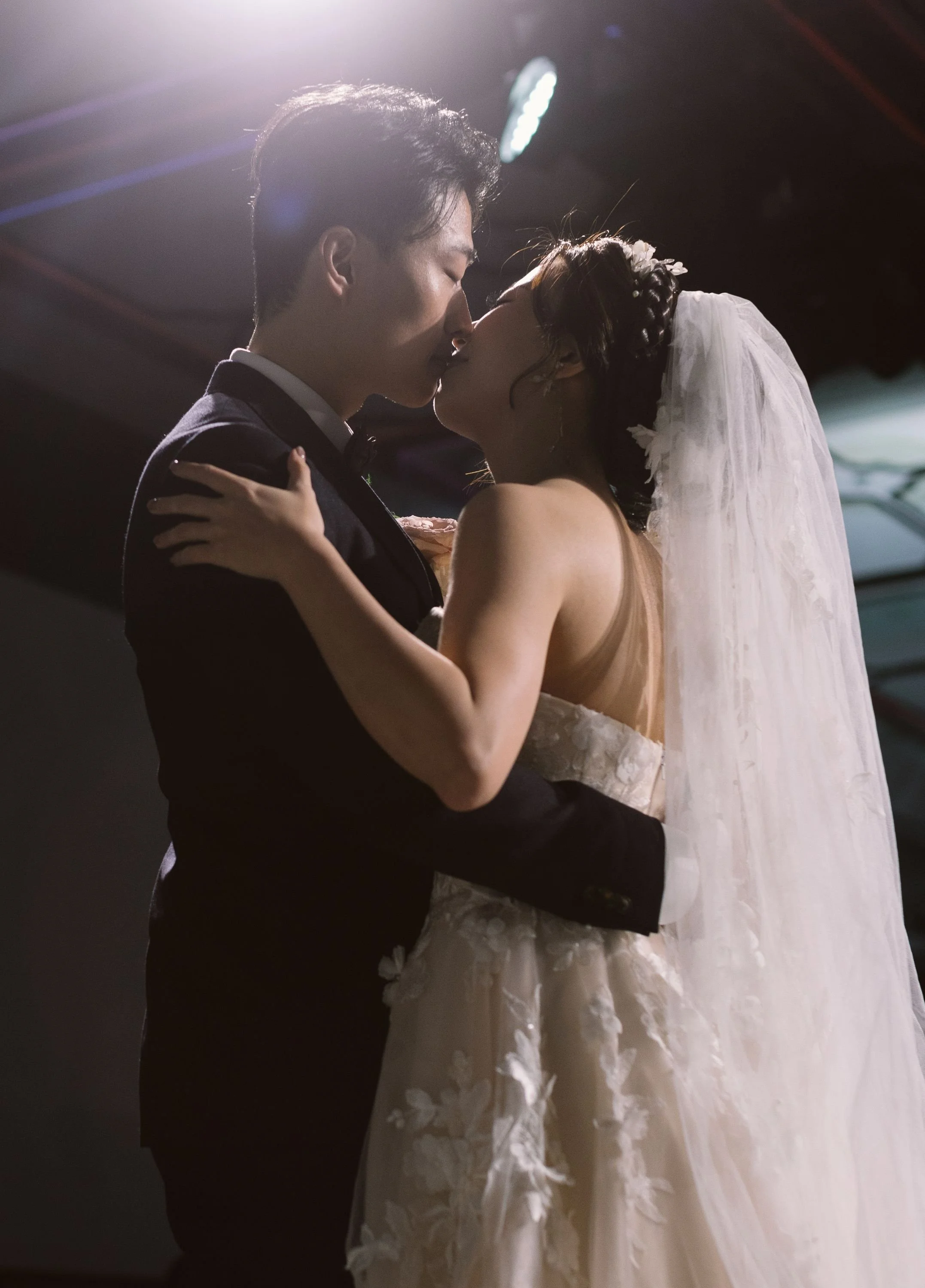 A bride and groom share a kiss during their wedding ceremony, with the bride wearing a white wedding dress and veil, and the groom in a black tuxedo.