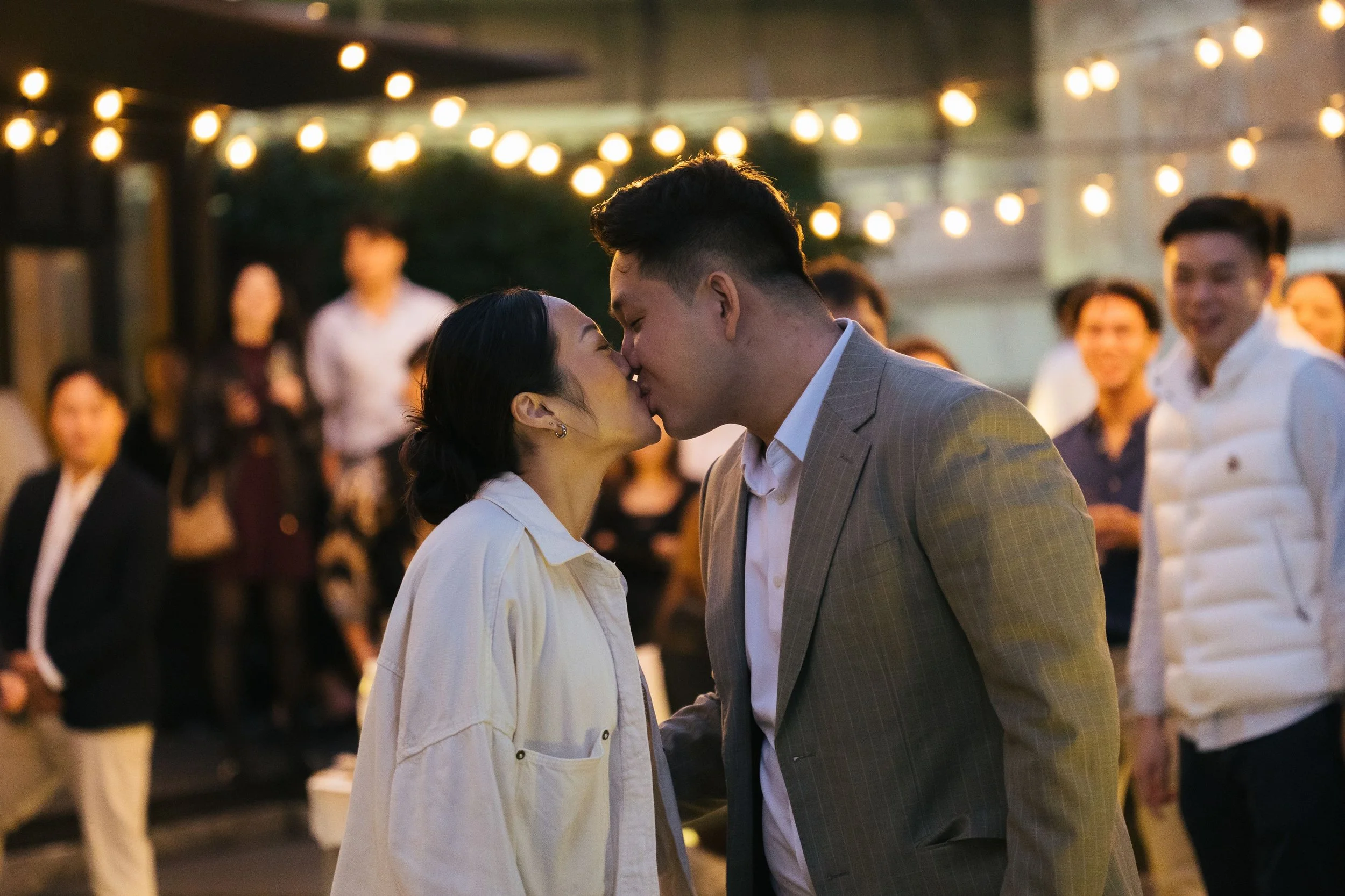 A couple kissing at night during an outdoor event, with string lights overhead and onlookers in the background.