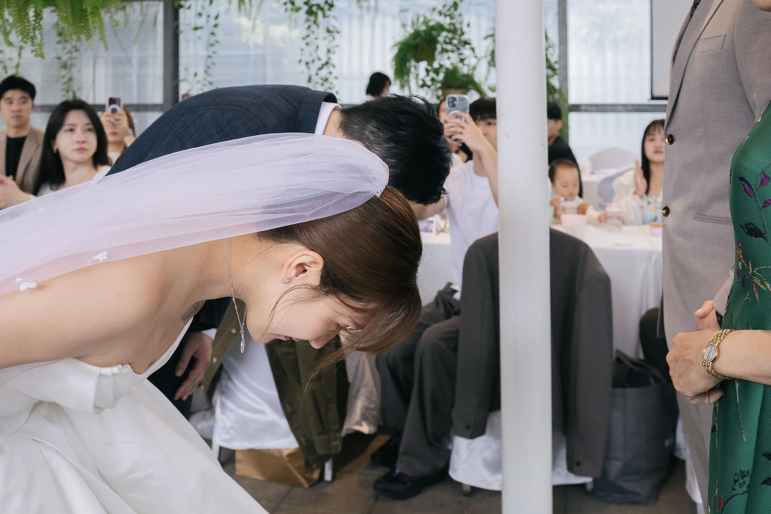 A woman in a wedding dress and a bride in a white gown bowing at a wedding ceremony, surrounded by seated guests taking photos.